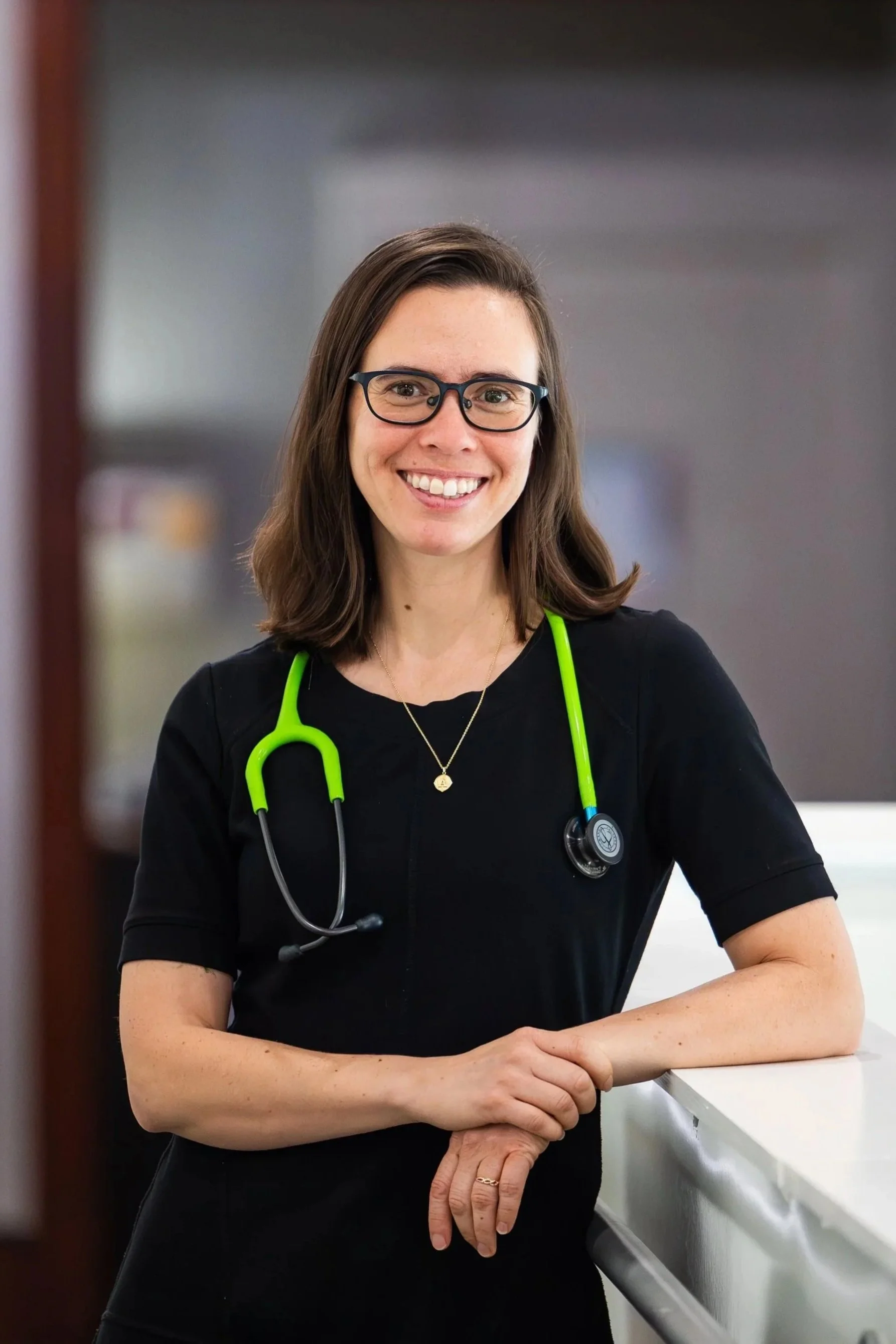 A smiling female healthcare professional with glasses, wearing a black uniform and a green stethoscope, standing at a hospital or clinic counter.