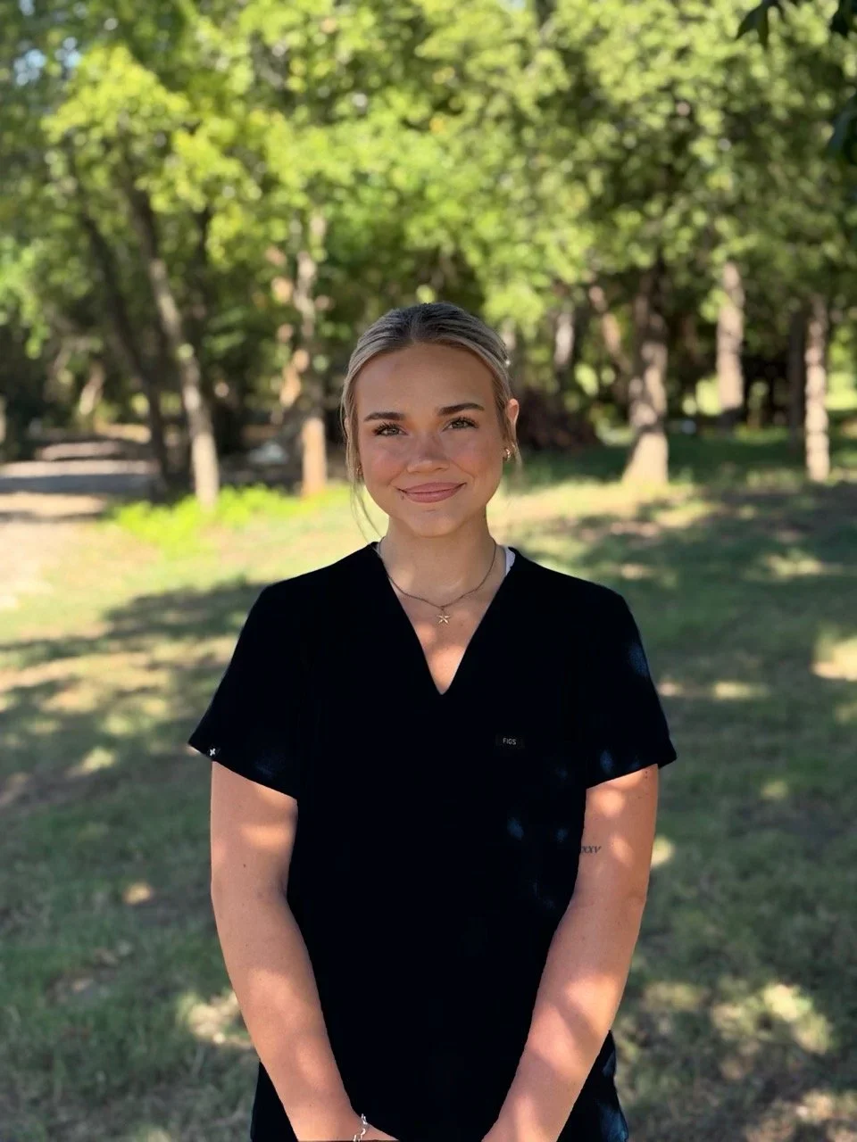 A young woman with blonde hair and fair skin standing outdoors in a park-like setting with trees and grass in the background. She is smiling and wearing a black V-neck shirt, small earrings, and a necklace.