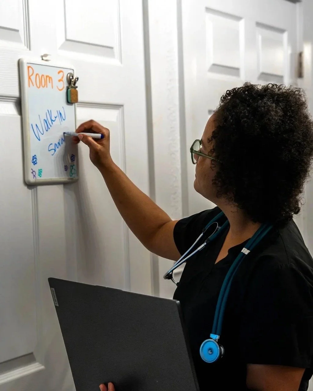 A healthcare worker, wearing glasses and a stethoscope, writes on a whiteboard that says 'Room 3' with the words 'Walking' and 'Sine' on it, located in a room with white paneled doors.