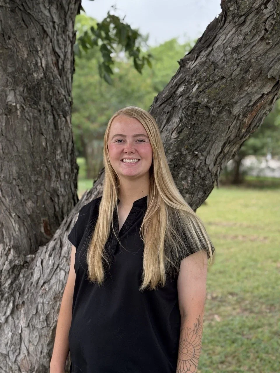 A young woman with long blonde hair, wearing a black shirt, smiling outdoors in front of a large tree with textured bark and green foliage.