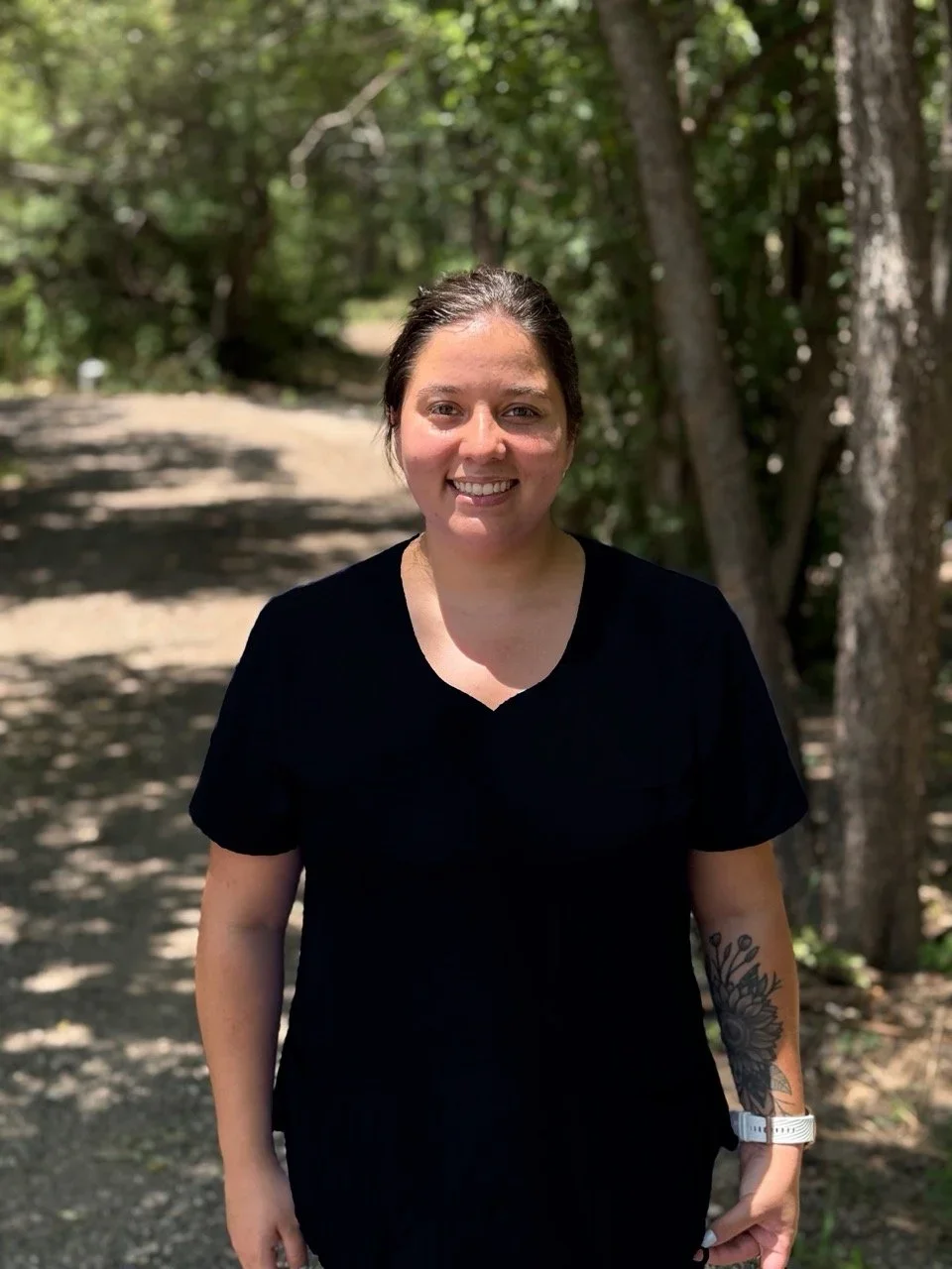A woman with dark hair tied back, smiling, standing outdoors on a sunny wooded trail, wearing a black shirt, with a tattoo of flowers on her left forearm and a white watch on her left wrist.