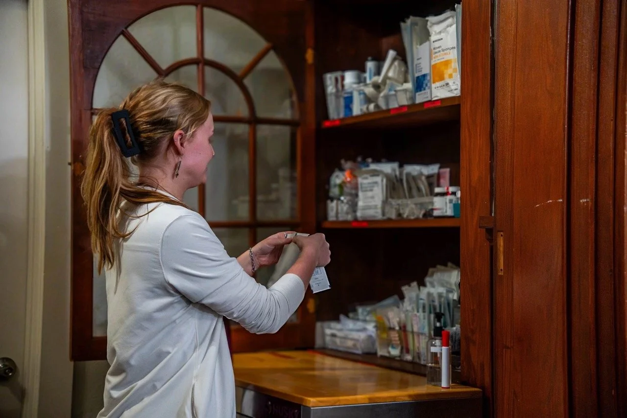 A woman with red hair and a black hair clip is holding a medicine bottle in front of shelves filled with various medical supplies in a wooden cabinet.