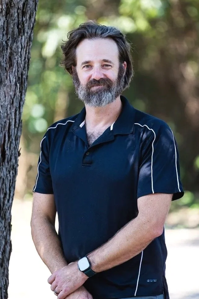 A man with a beard and shoulder-length brown hair standing outdoors near a tree with a blurred background of foliage. He is wearing a navy blue polo shirt and a black wristwatch.