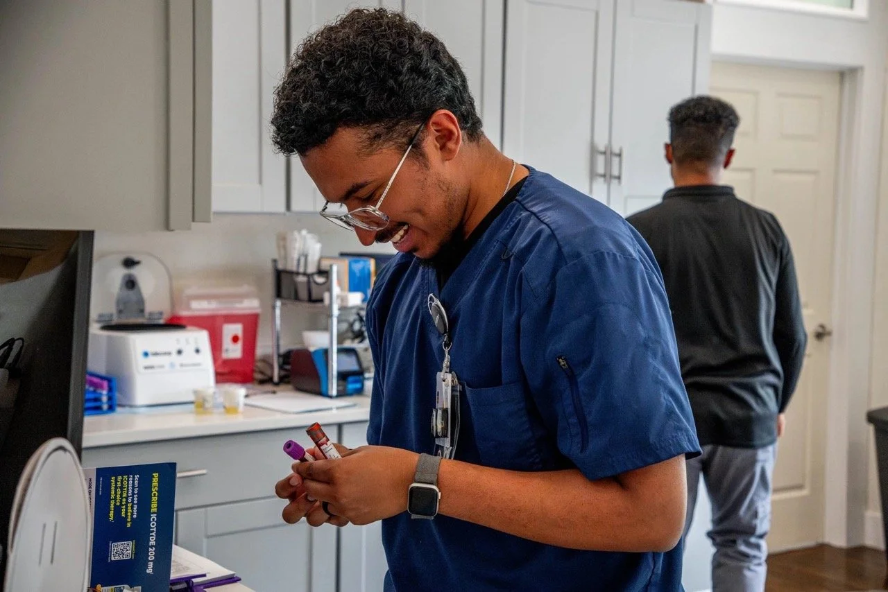 A smiling healthcare worker in blue scrubs holding and inspecting test tubes in a medical clinic or laboratory.