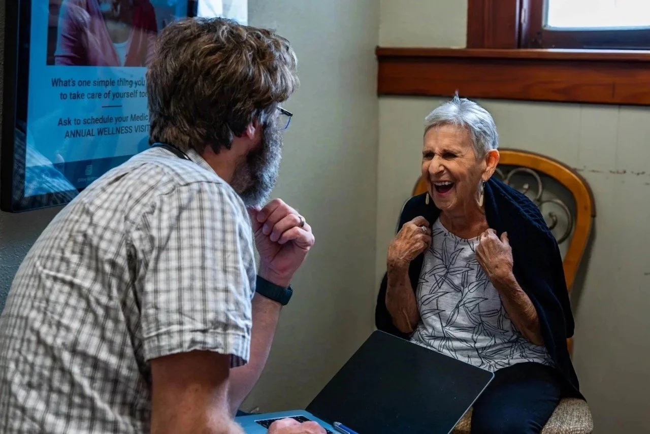 Dr. Mahaffey is sitting having an engaged conversation with an elderly patient. The patient is an elderly woman with short grey hair, and appears to be laughing with a big smile.