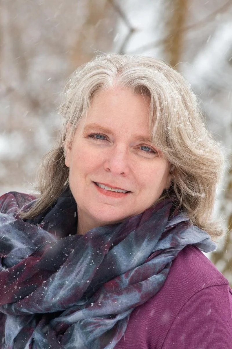 A woman with blonde hair and blue eyes smiling outdoors in a snowy setting, wearing a maroon top and a dark, patterned scarf.