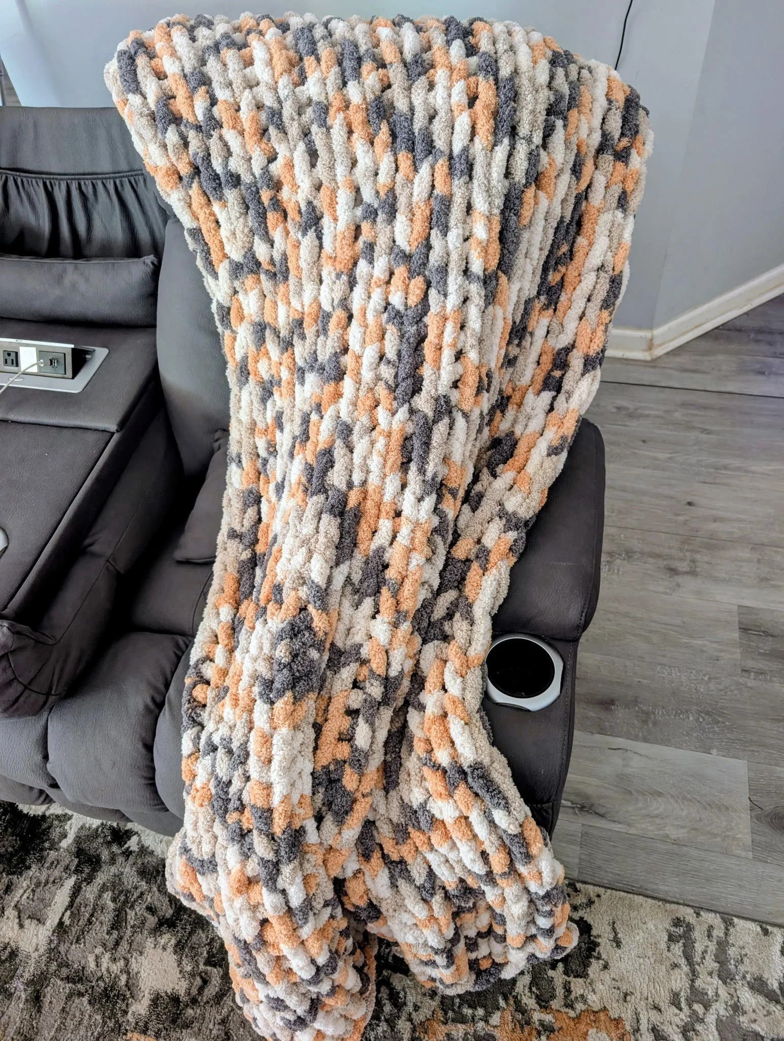 A cozy multicolored knit blanket,  in beige, orange, gray, and white draped over the back of a gray sofa in a living room with gray wood flooring, a patterned rug, and a small circular side table.