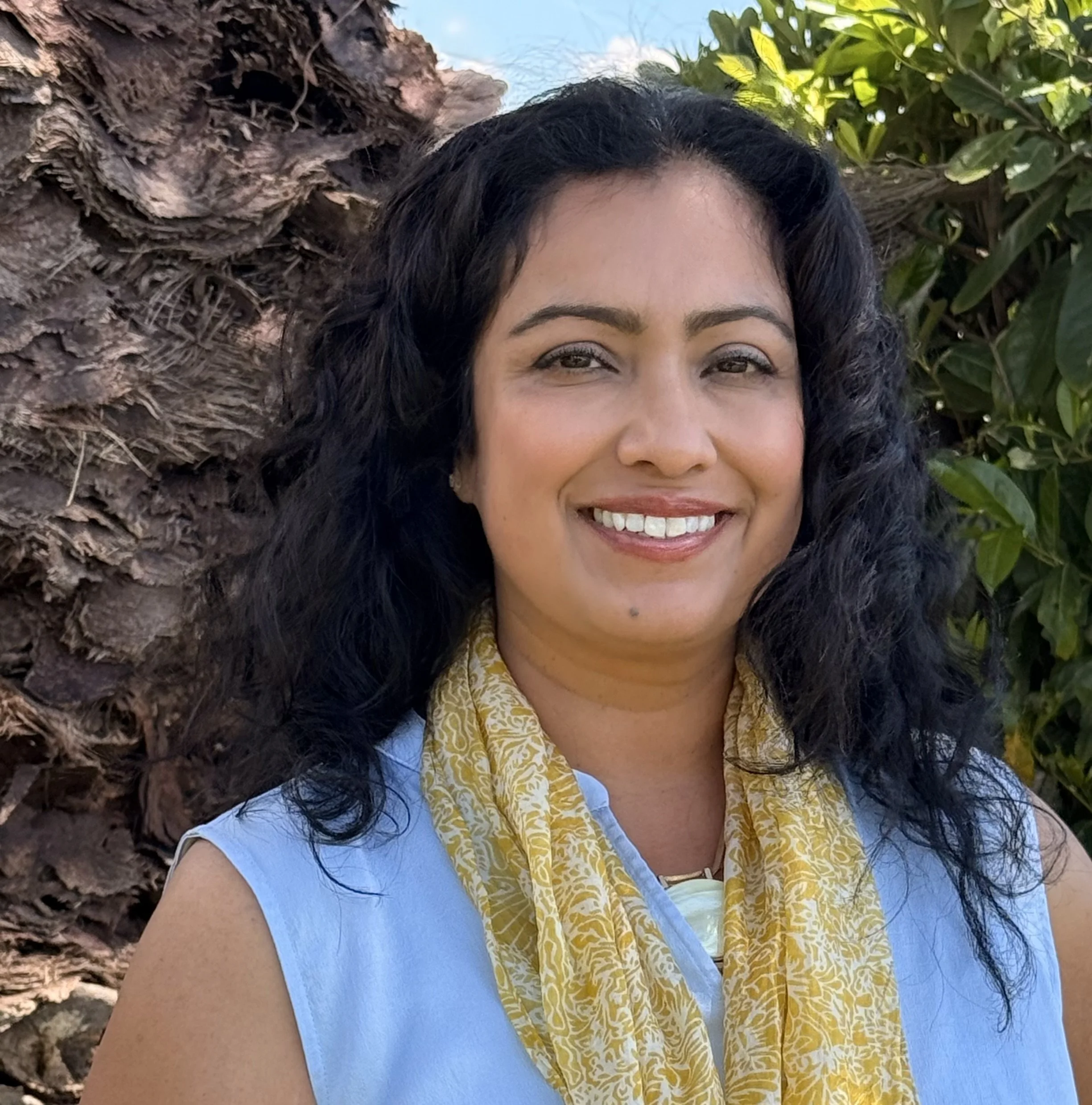 A woman with dark, curly hair smiling outdoors with a blue sky, a tree trunk, and green leaves in the background.
