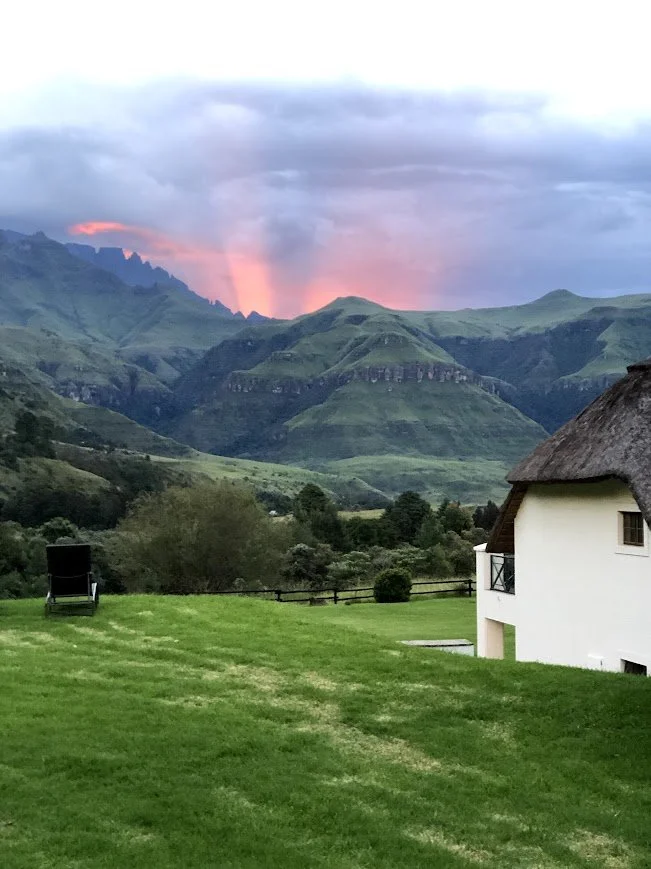 Scenic view of green rolling hills and mountains at sunset with a pink and purple sky, a thatched-roof house, and a lawn chair in the foreground.