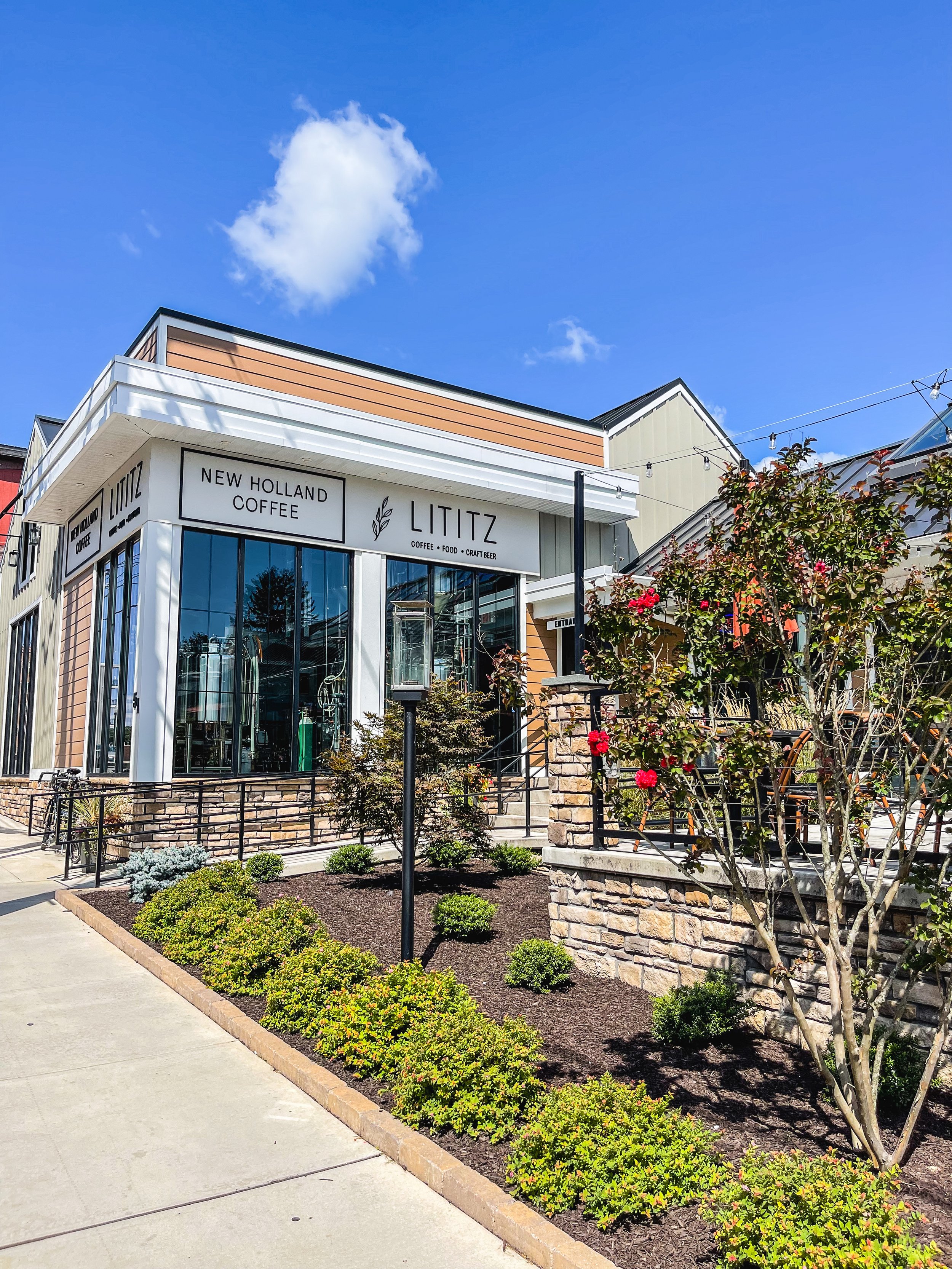 Exterior of a modern coffee shop named 'Lititz' with large glass windows, a sign indicating 'New Holland Coffee,' and a well-maintained sidewalk and landscaping with bushes, small trees, and flowers under a blue sky with a few clouds.