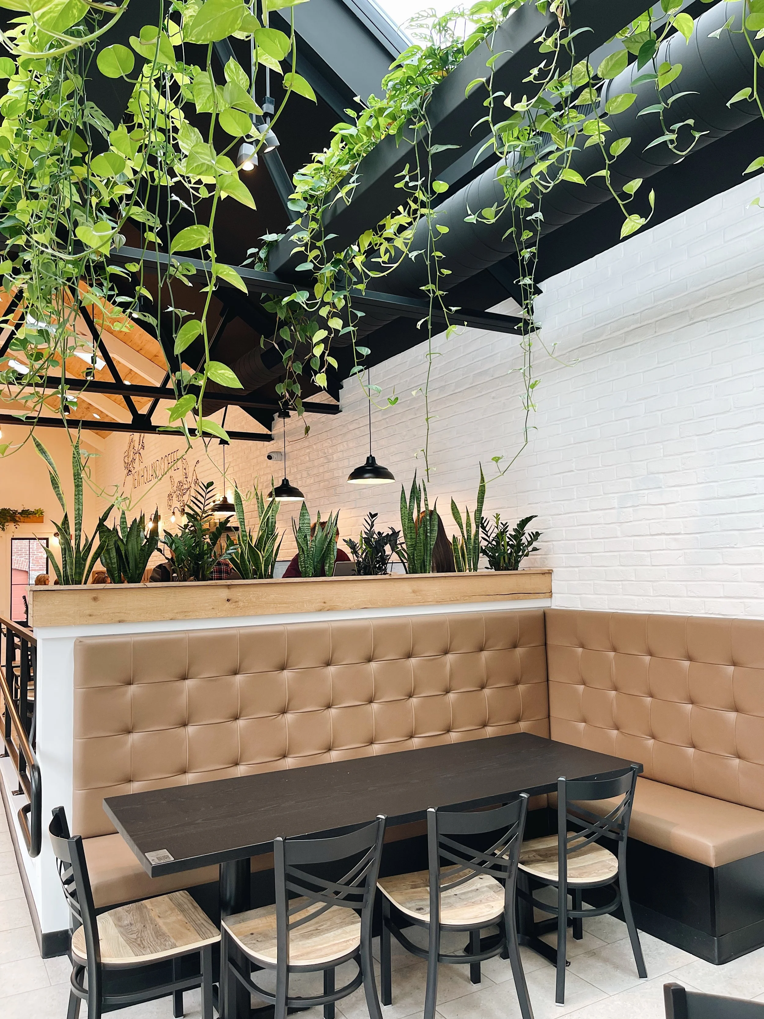 Indoor seating area with beige padded bench against white brick wall, black tables, black chairs with wooden seats, green plants on a partition, and hanging black pendant lights.