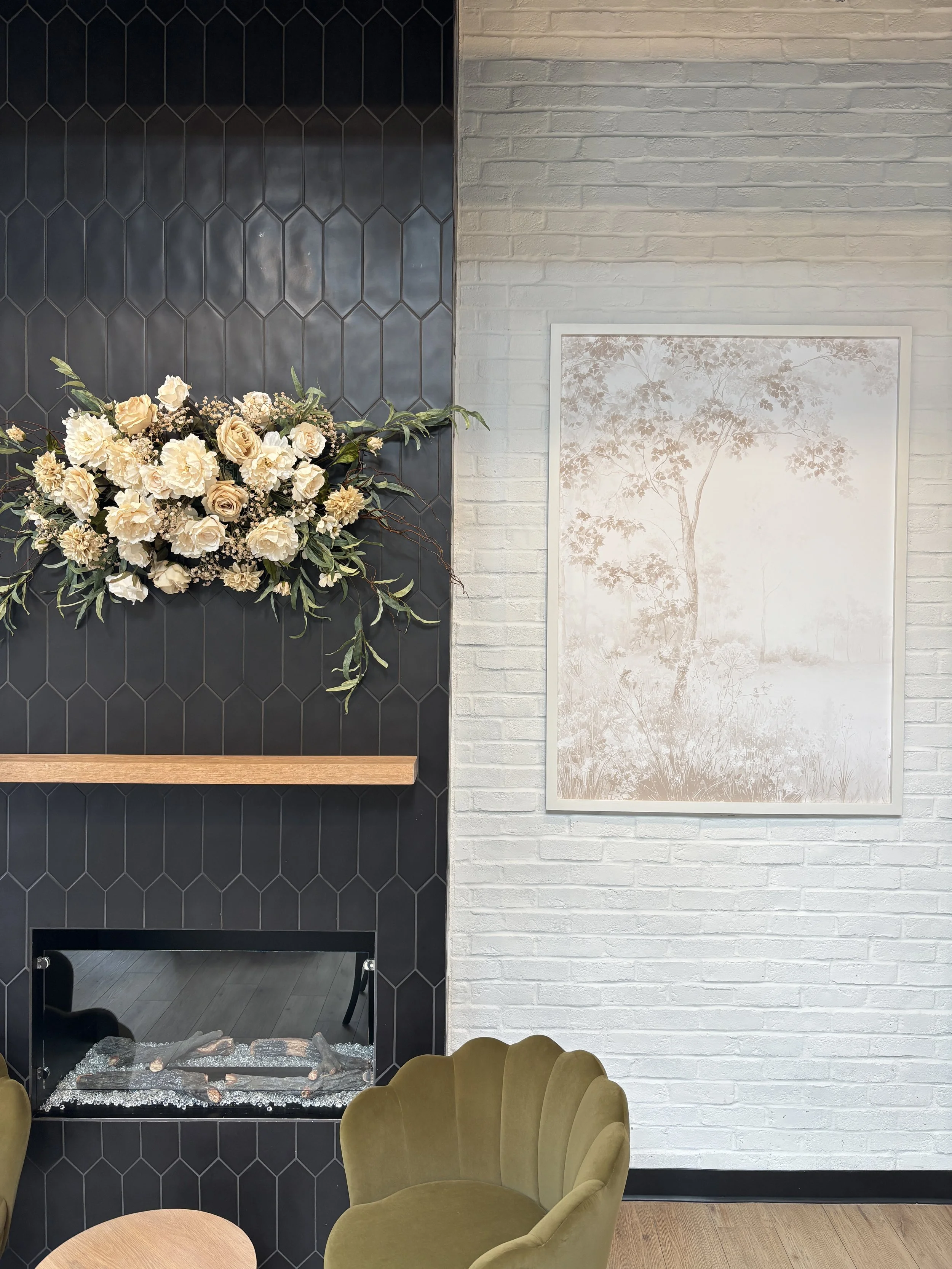 Indoor scene with a black tiled fireplace with metal accents, a bouquet of cream-colored roses and greenery on the mantel, a framed monochrome tree print on a white brick wall, a green upholstered chair with a scalloped backrest in the foreground, a small round wooden table, and wooden flooring.