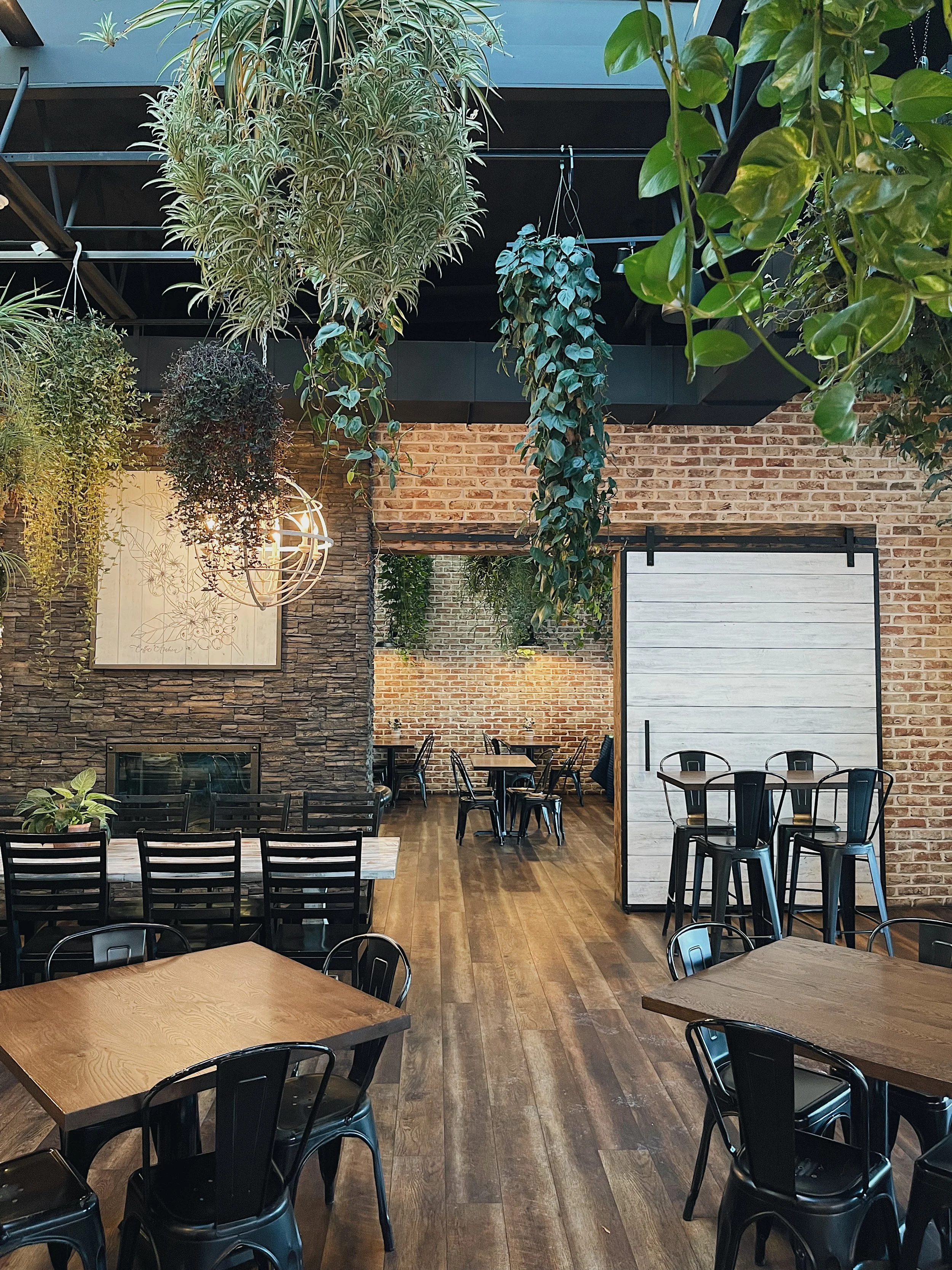 Interior of a modern, rustic-style restaurant or cafe with wooden tables, black chairs, hanging plants, and brick walls.