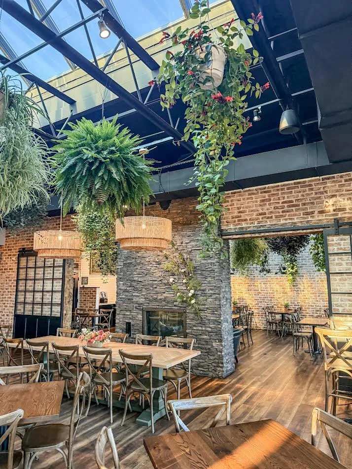 Interior of a restaurant with wooden tables and chairs, a stone fireplace, hanging plants, brick walls, and large windows letting in natural light.
