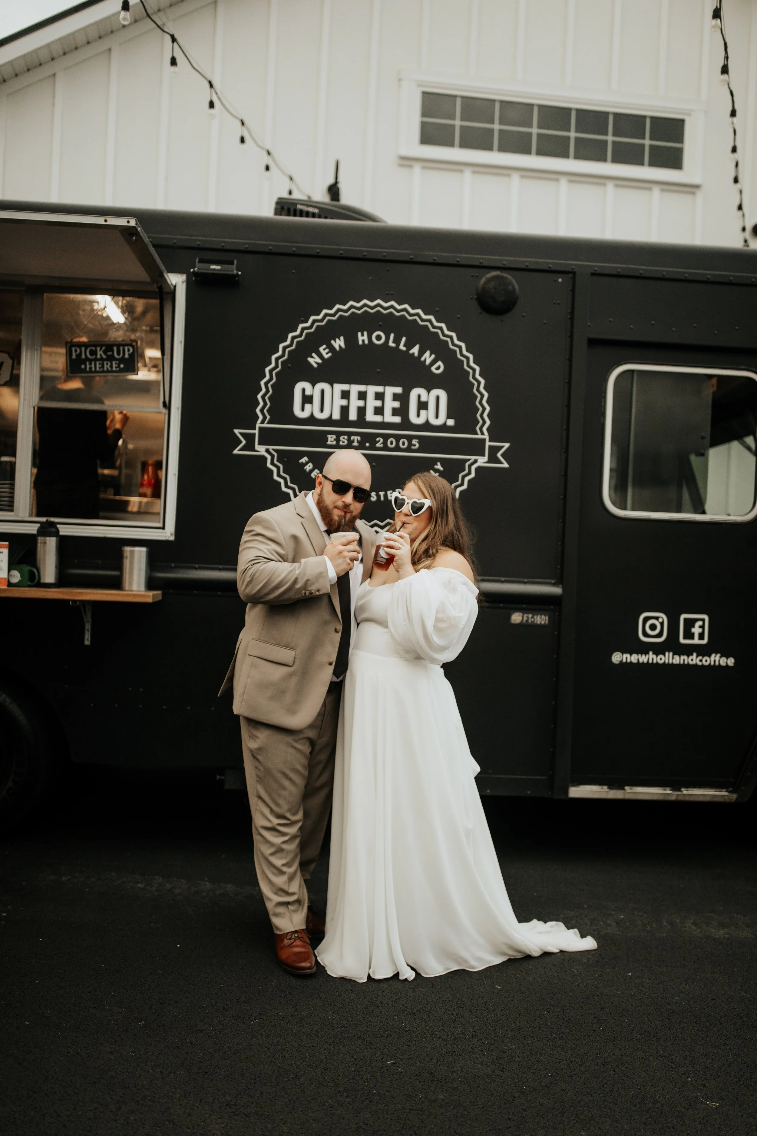 A bride and groom standing in front of a black coffee truck, sharing a drink and smiling.
