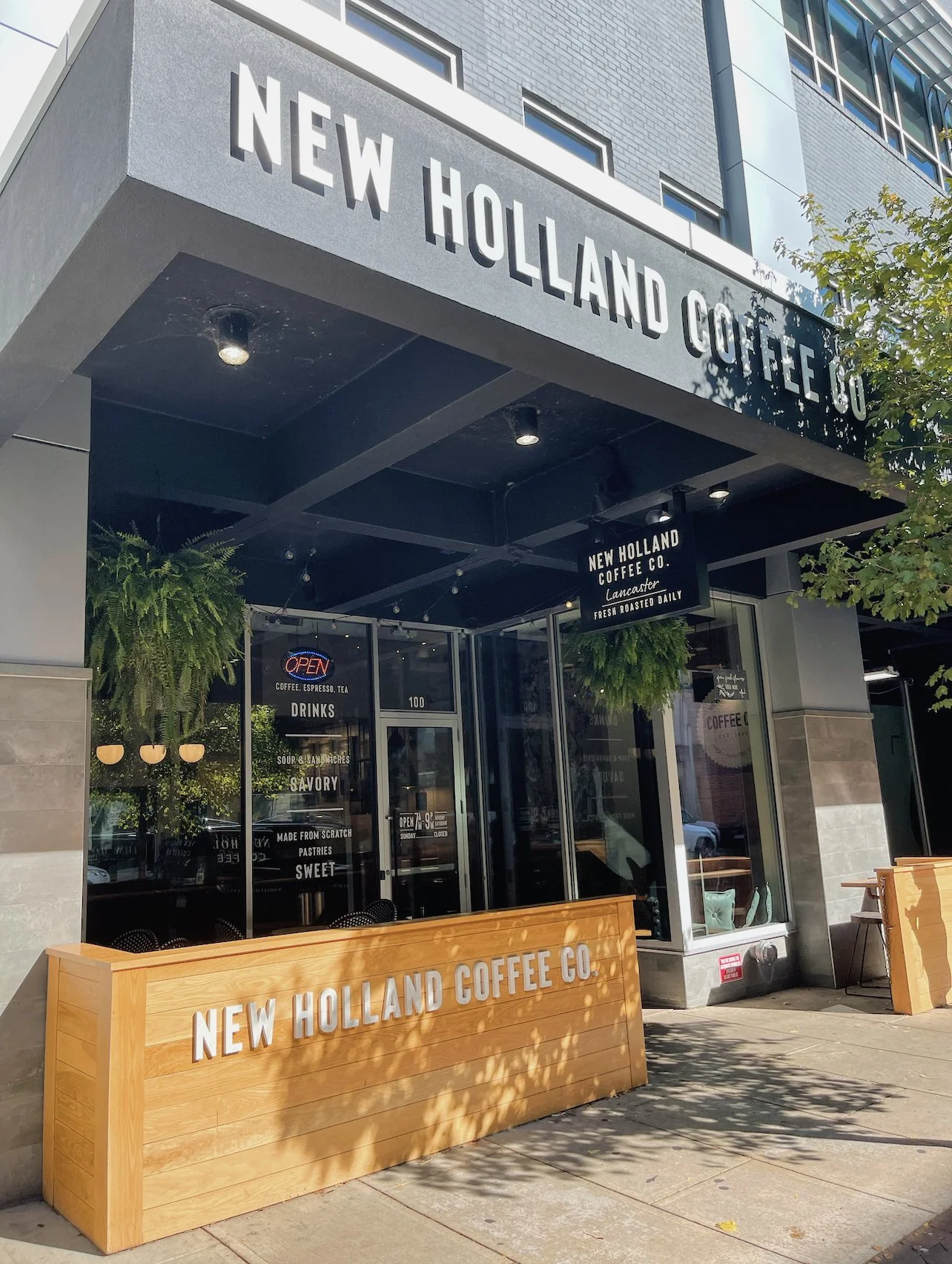 Exterior view of a modern coffee shop called New Holland Coffee Co. with outdoor seating, potted plants, and signage indicating it serves coffee, espresso, tea, drinks, and baked goods.