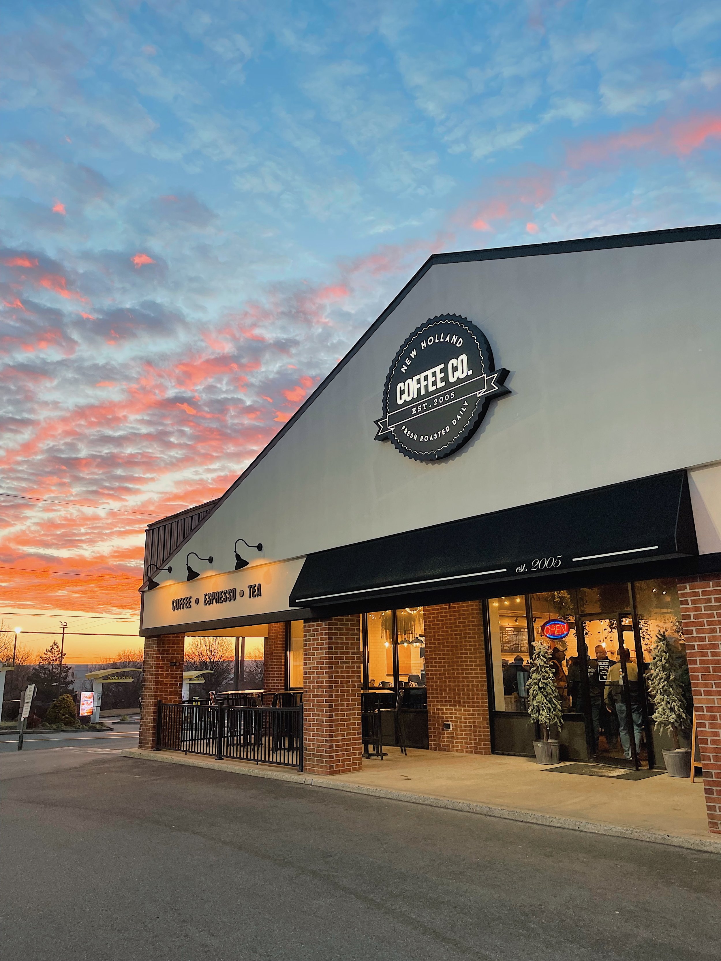 Exterior view of a coffee shop named 'New Holland Coffee Co.' with a sunset sky in the background. The storefront has brick columns, large windows, and a sign indicating it was established in 2005. There are customers inside and a neon 'Open' sign in the window.