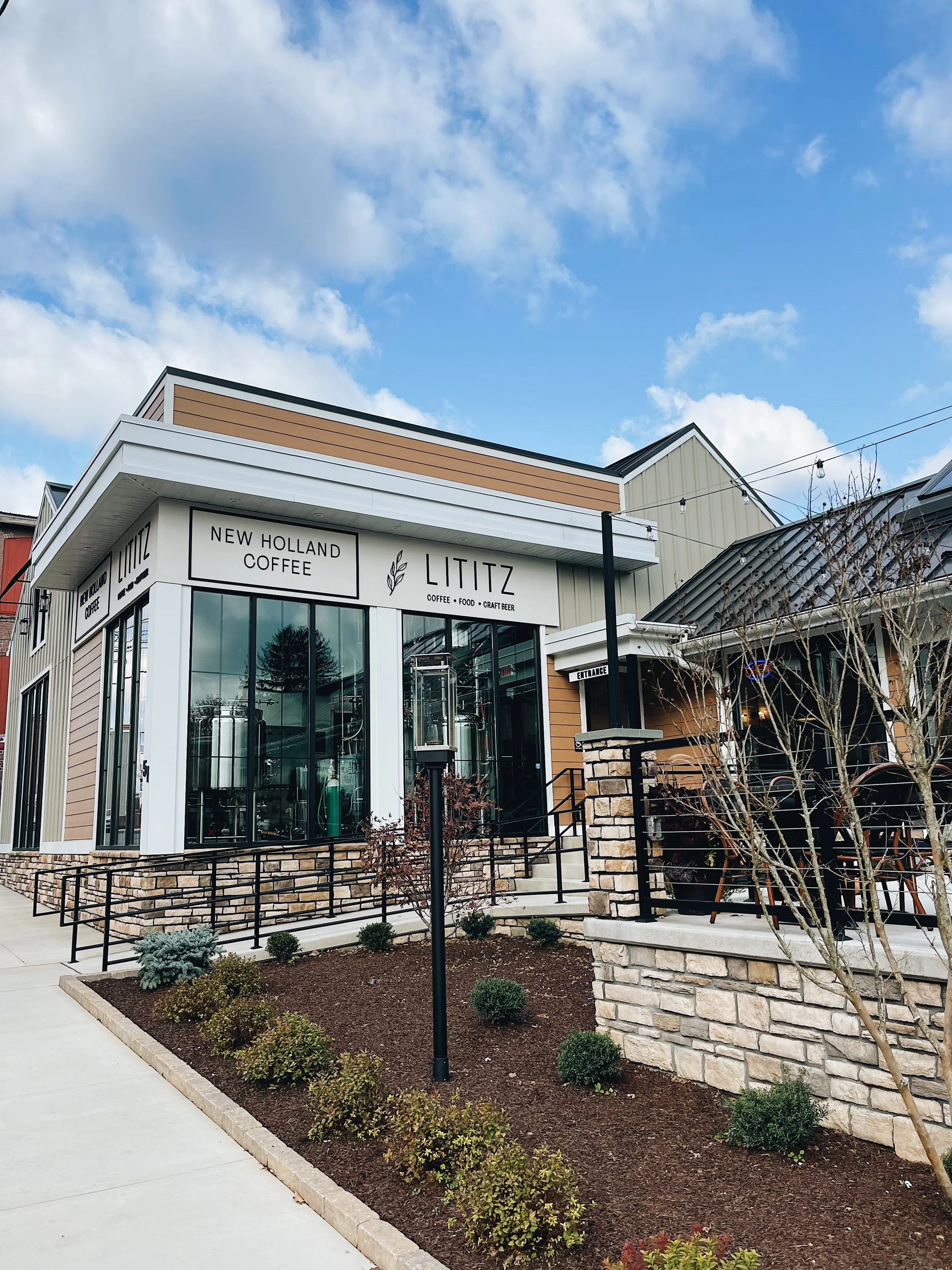 Exterior view of a modern coffee shop called 'Lititz' with signage indicating it offers coffee, food, and craft beer. The building has a combination of beige and white siding, large glass windows, and a stone foundation. There are landscaped plants and a sidewalk in front, with string lights hanging overhead against a partly cloudy sky.