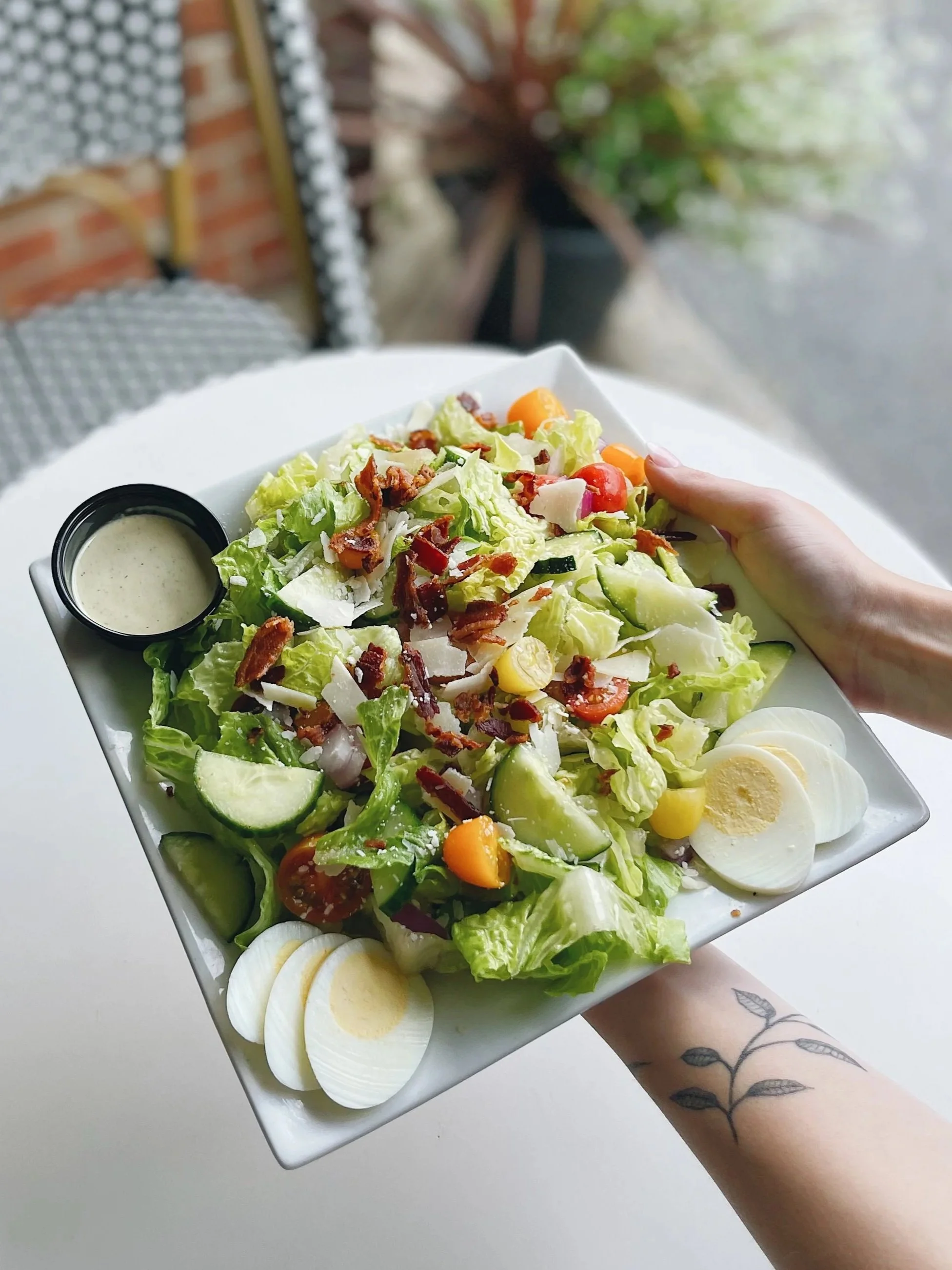 A hand holding a rectangular white plate filled with a fresh salad that contains lettuce, cherry tomatoes, cucumber, shredded cheese, bacon bits, and sliced boiled eggs, with a small cup of dressing on the side, against a blurred background.