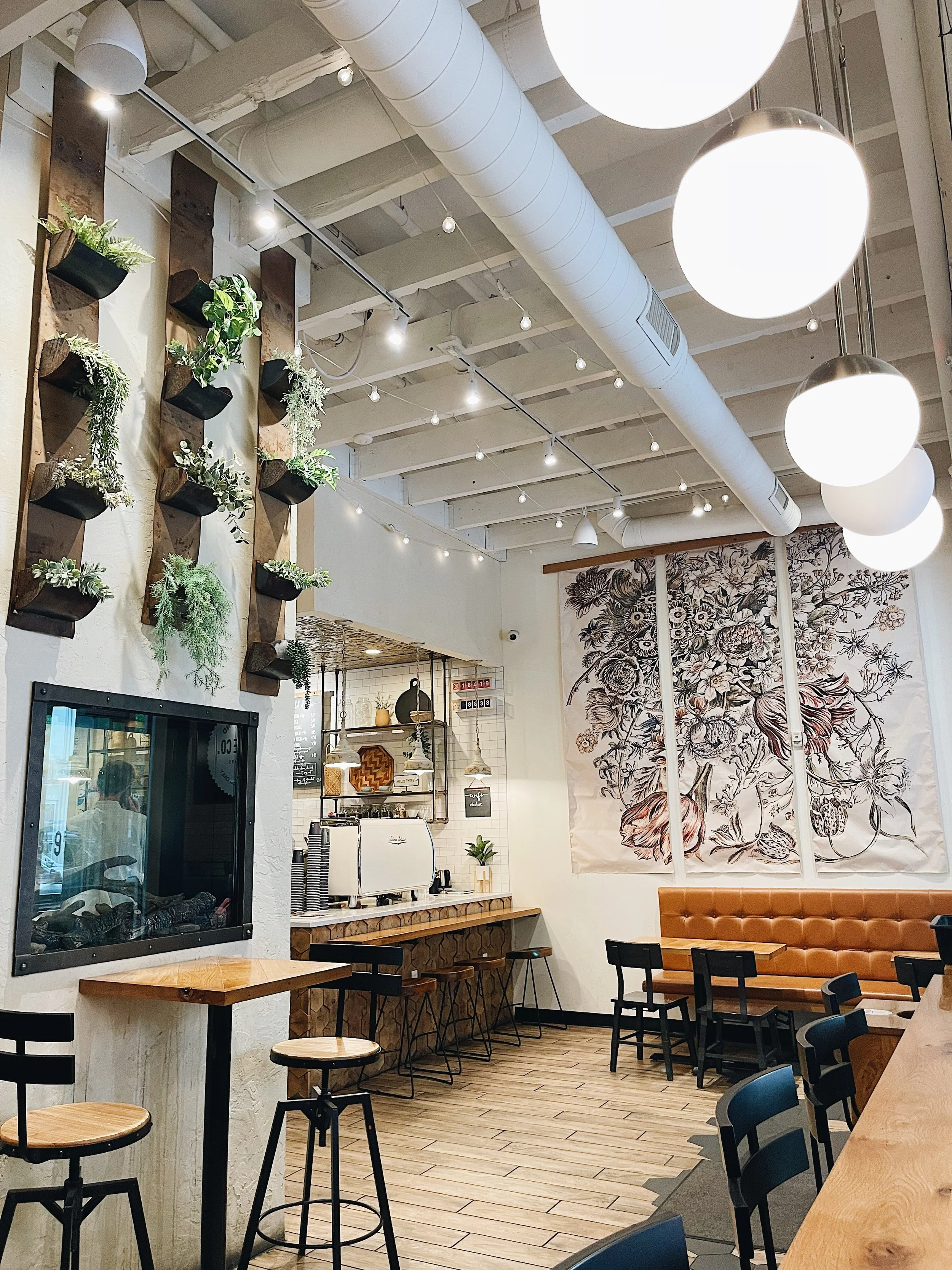 Interior of a modern cafe with hanging lights, wooden furniture, potted plants on the wall, and a large floral mural on the wall.