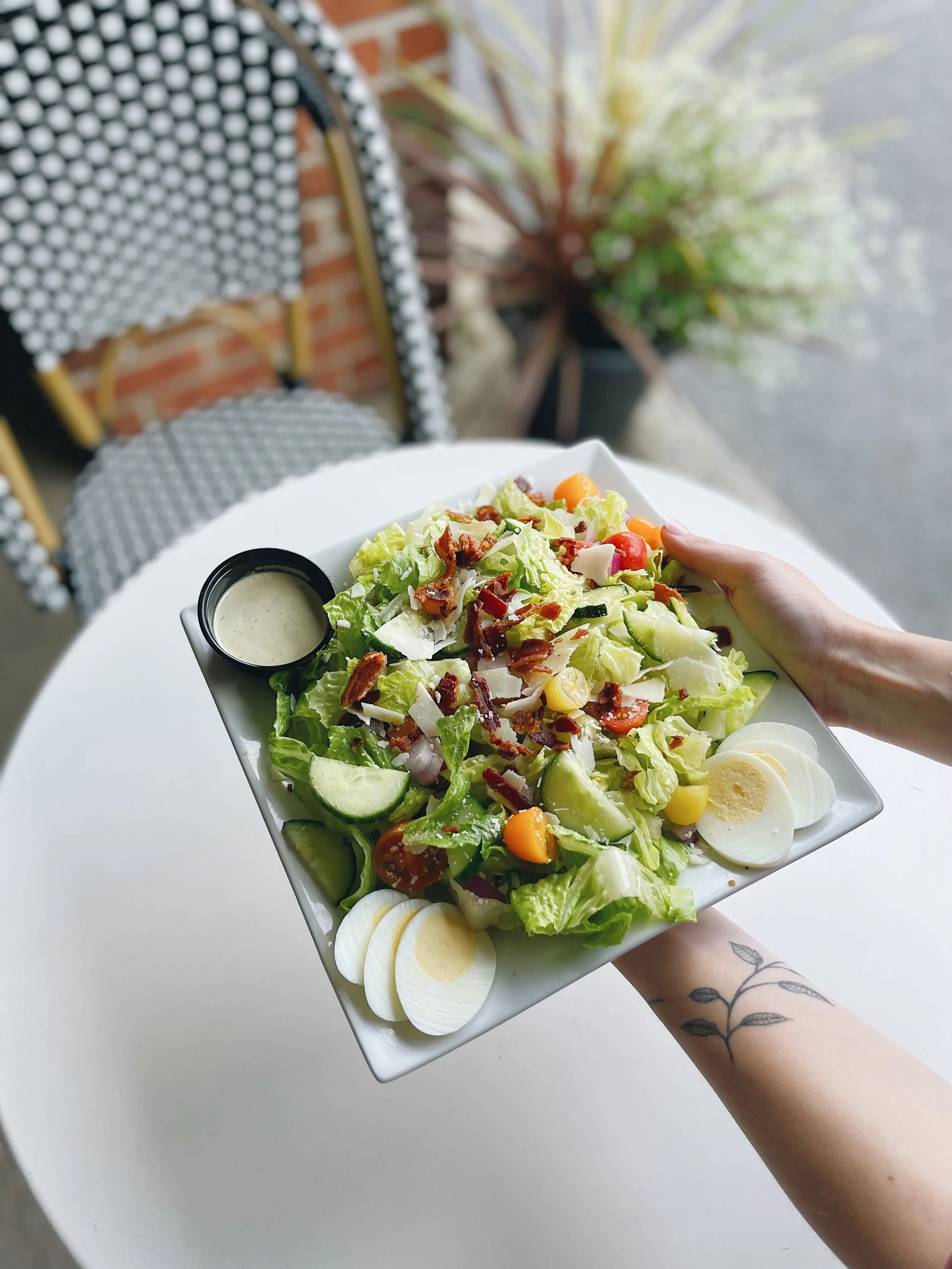 Person holding a rectangular white plate with a fresh garden salad, sliced hard-boiled eggs, and a small container of dressing, sitting at a white table outdoors with a chair and plant in the background.