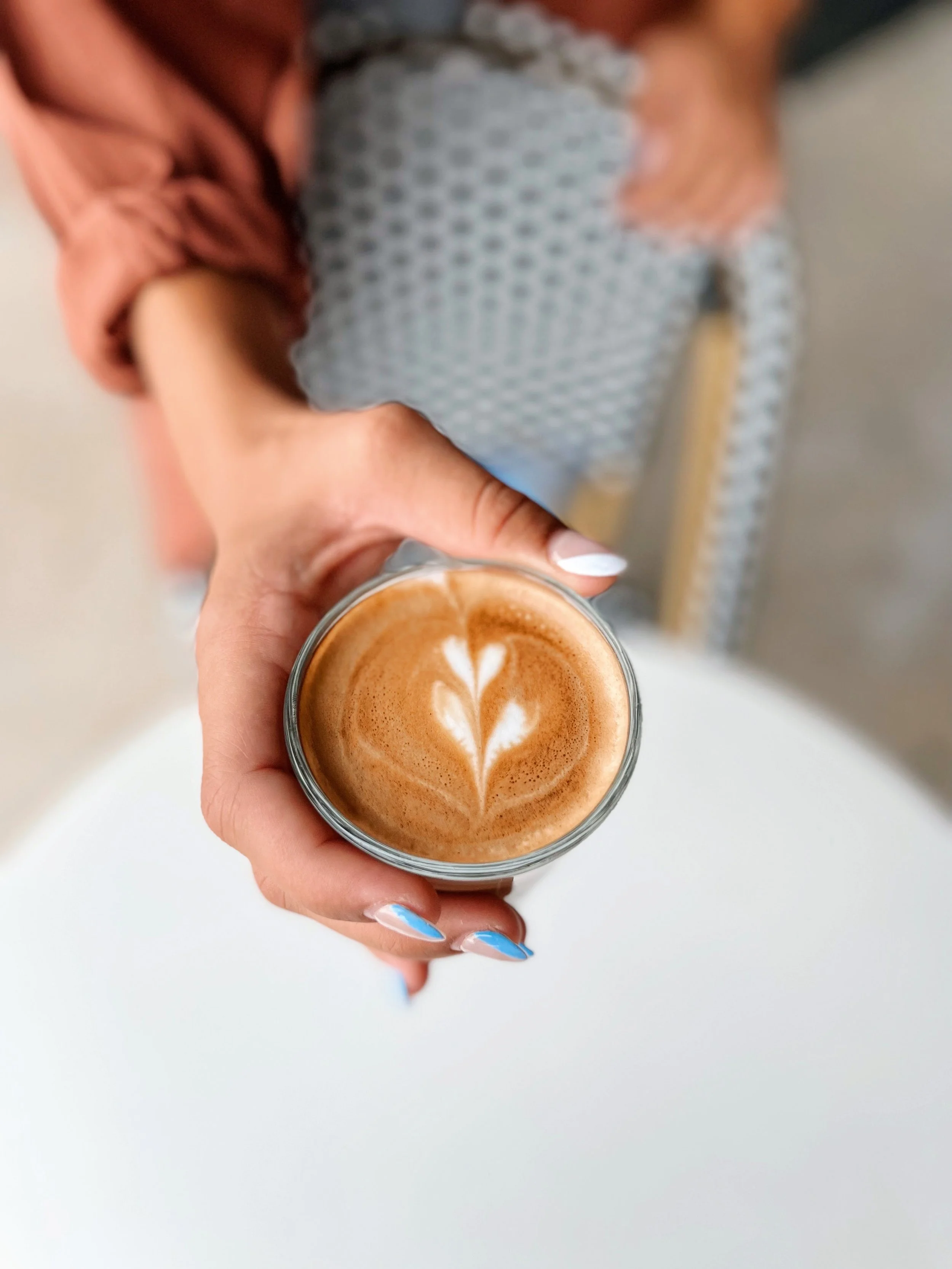 A person's hand with painted nails holding a glass of latte with foam art on top. Background shows a person's lap in a checkered skirt and a pink jacket.