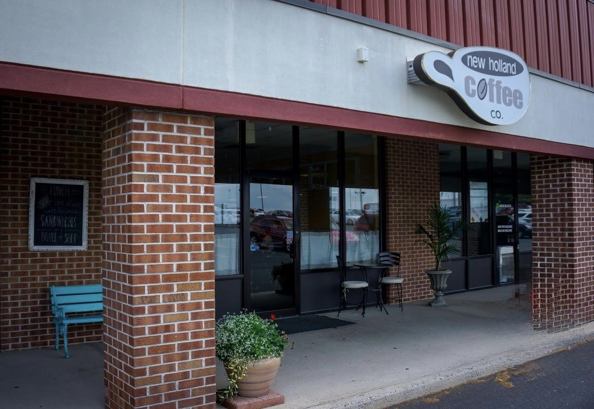 Exterior of a coffee shop named 'New Holland Coffee Co.' with large glass windows, brick columns, outdoor seating with chairs and potted plants, situated in a shopping plaza.
