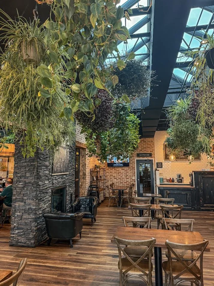 Interior of a modern restaurant or cafe with hanging plants, a stone fireplace, wooden tables and chairs, and a brick wall.
