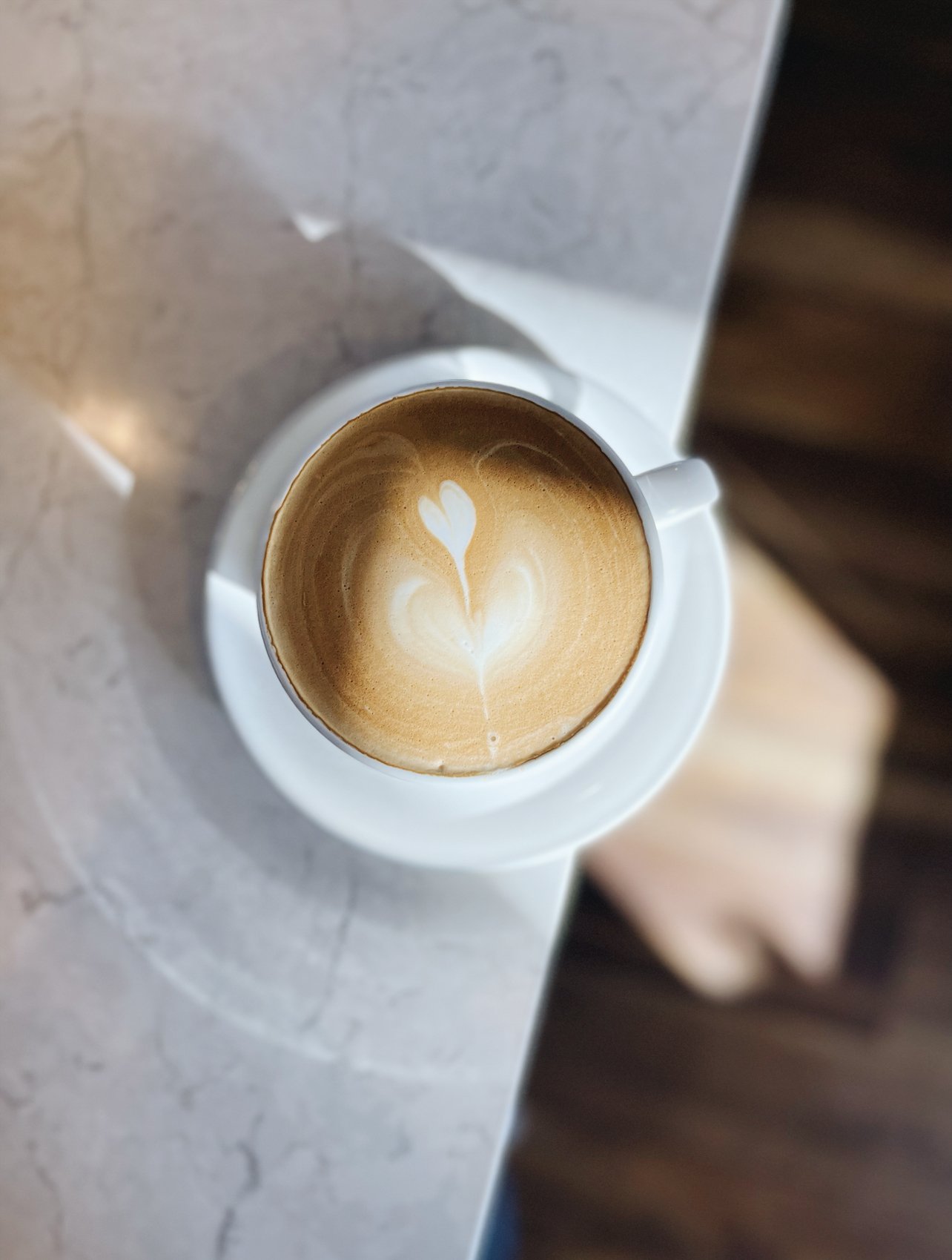 A white coffee cup with latte art in the shape of a heart, placed on a saucer on a marble countertop.