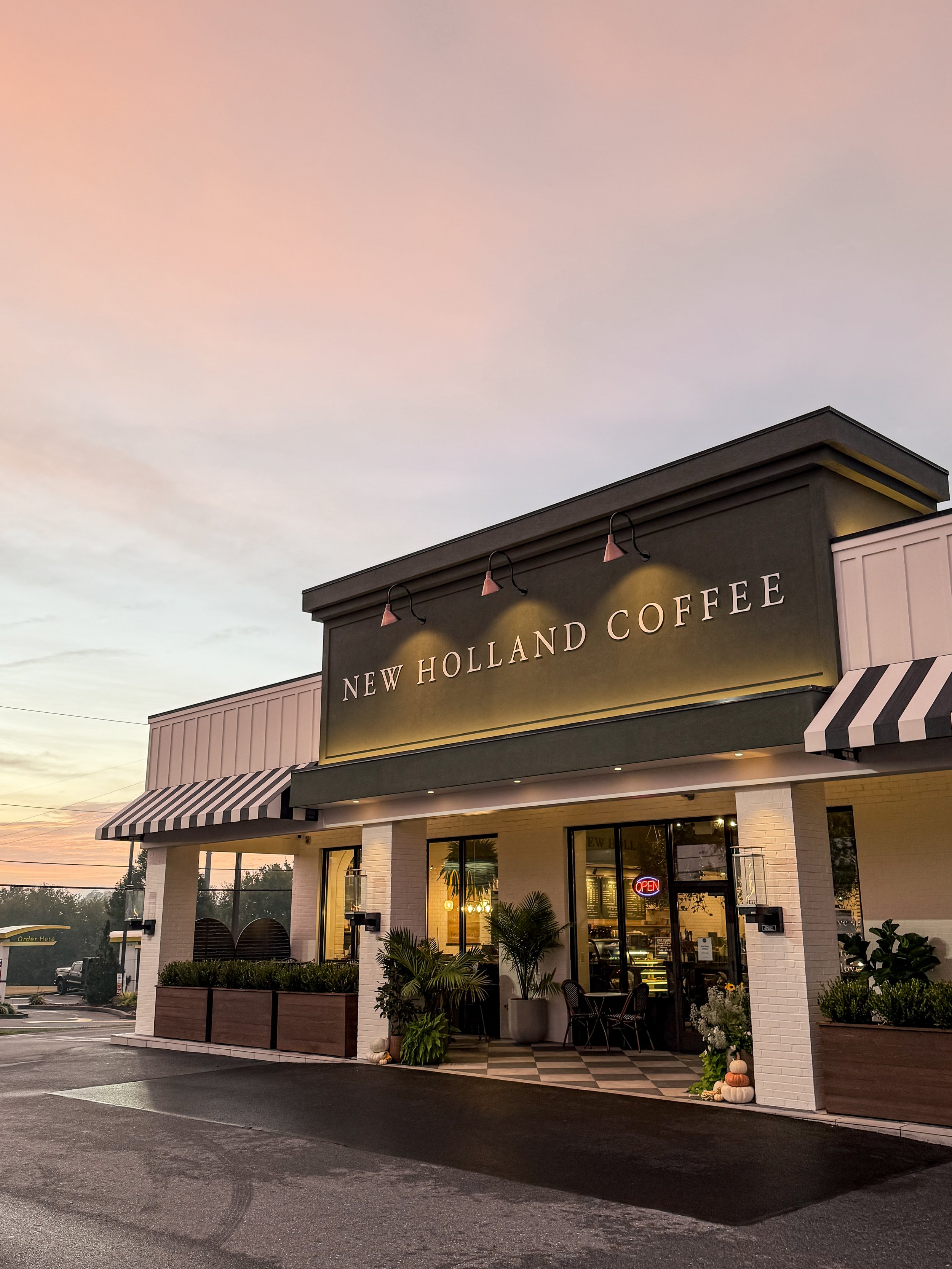 Exterior of a coffee shop with a sign that reads 'New Holland Coffee', sunset sky in the background, outdoor seating with plants, and autumn decorations at the entrance.