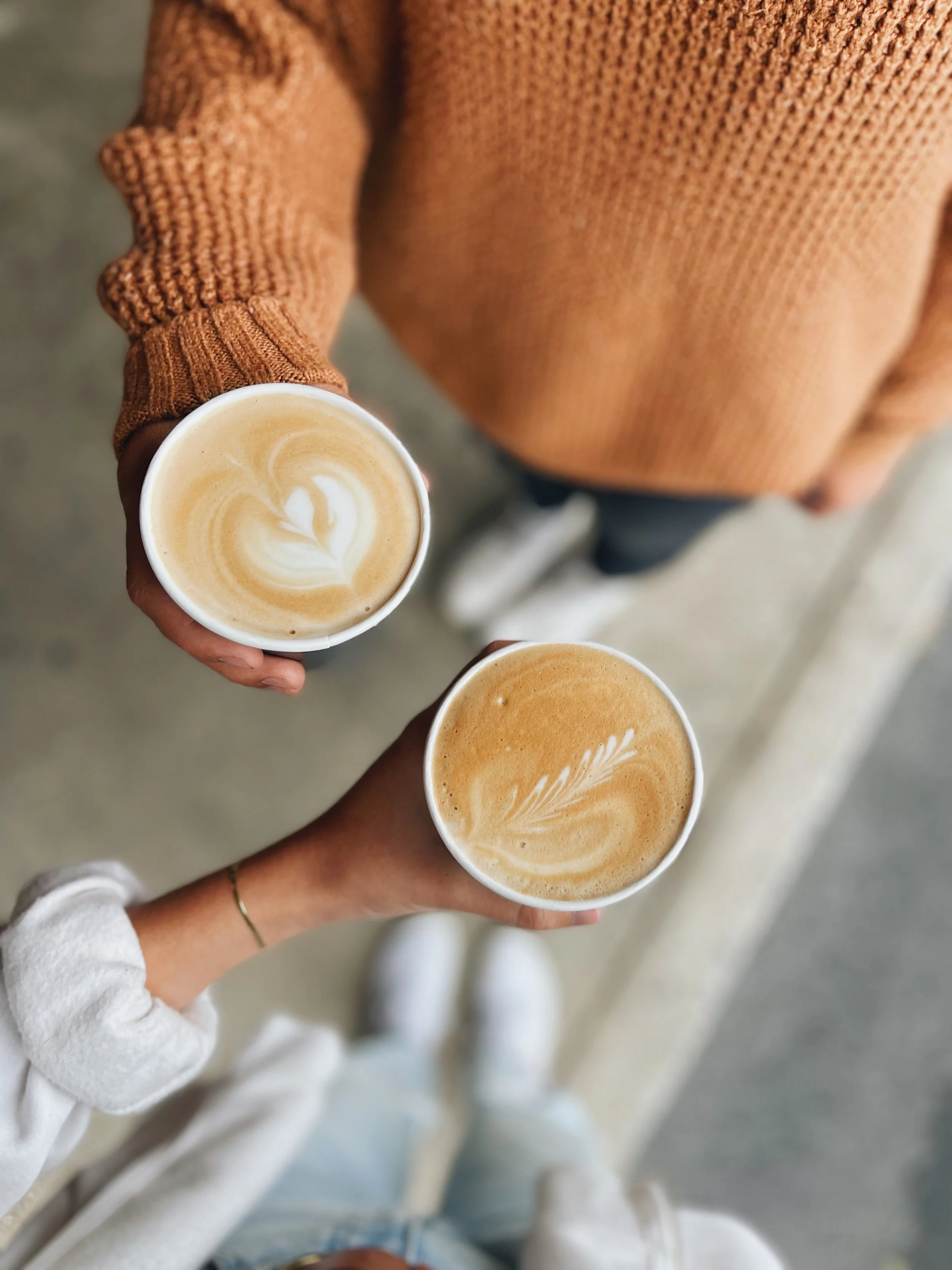Two hands holding cups of coffee with latte art, one with a gold bracelet, one wearing a white sweater, background shows a person in a brown sweater.