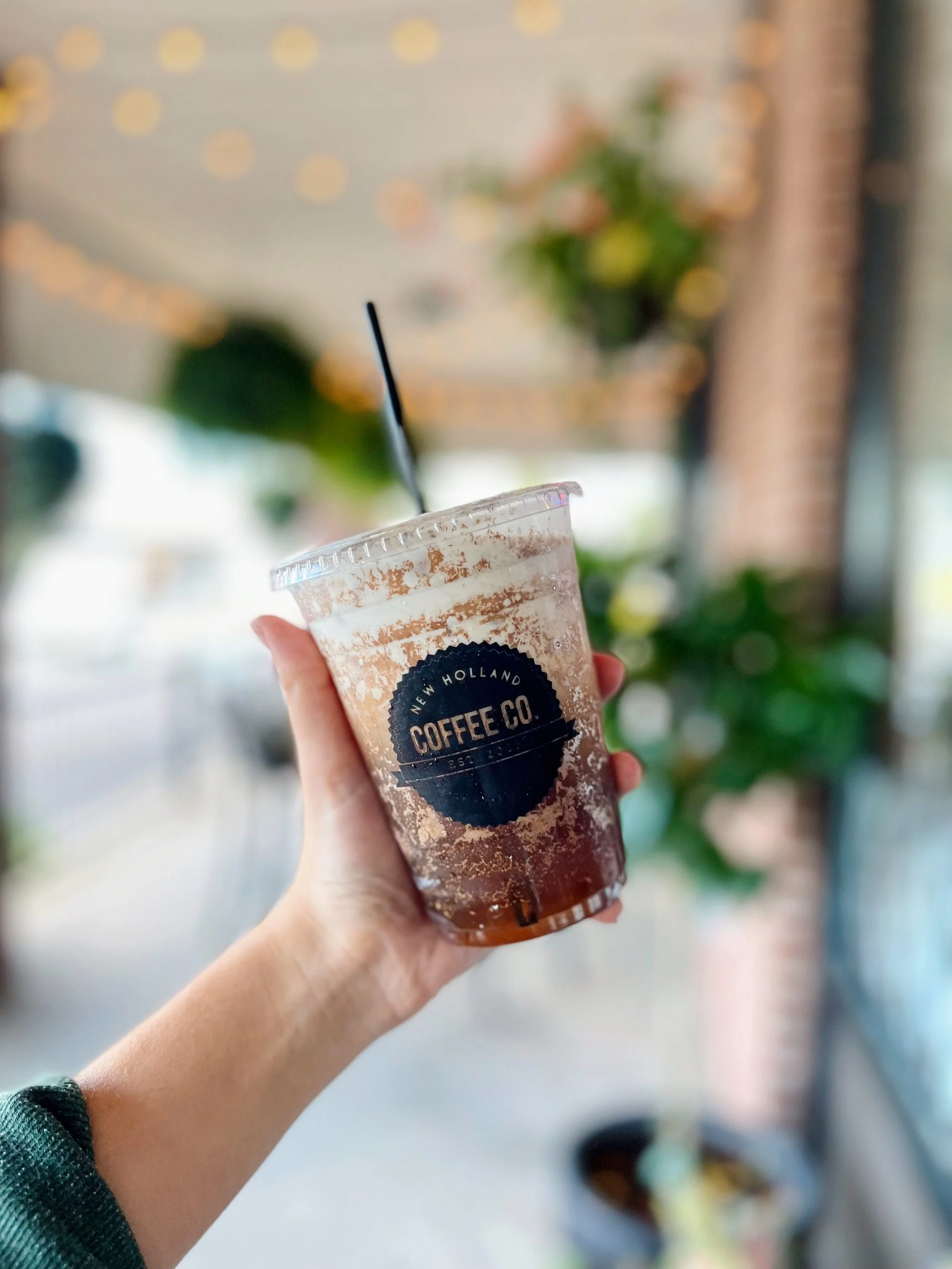 Person holding a plastic cup of iced coffee with a black straw in front of blurred background of Christmas lights and a plant.