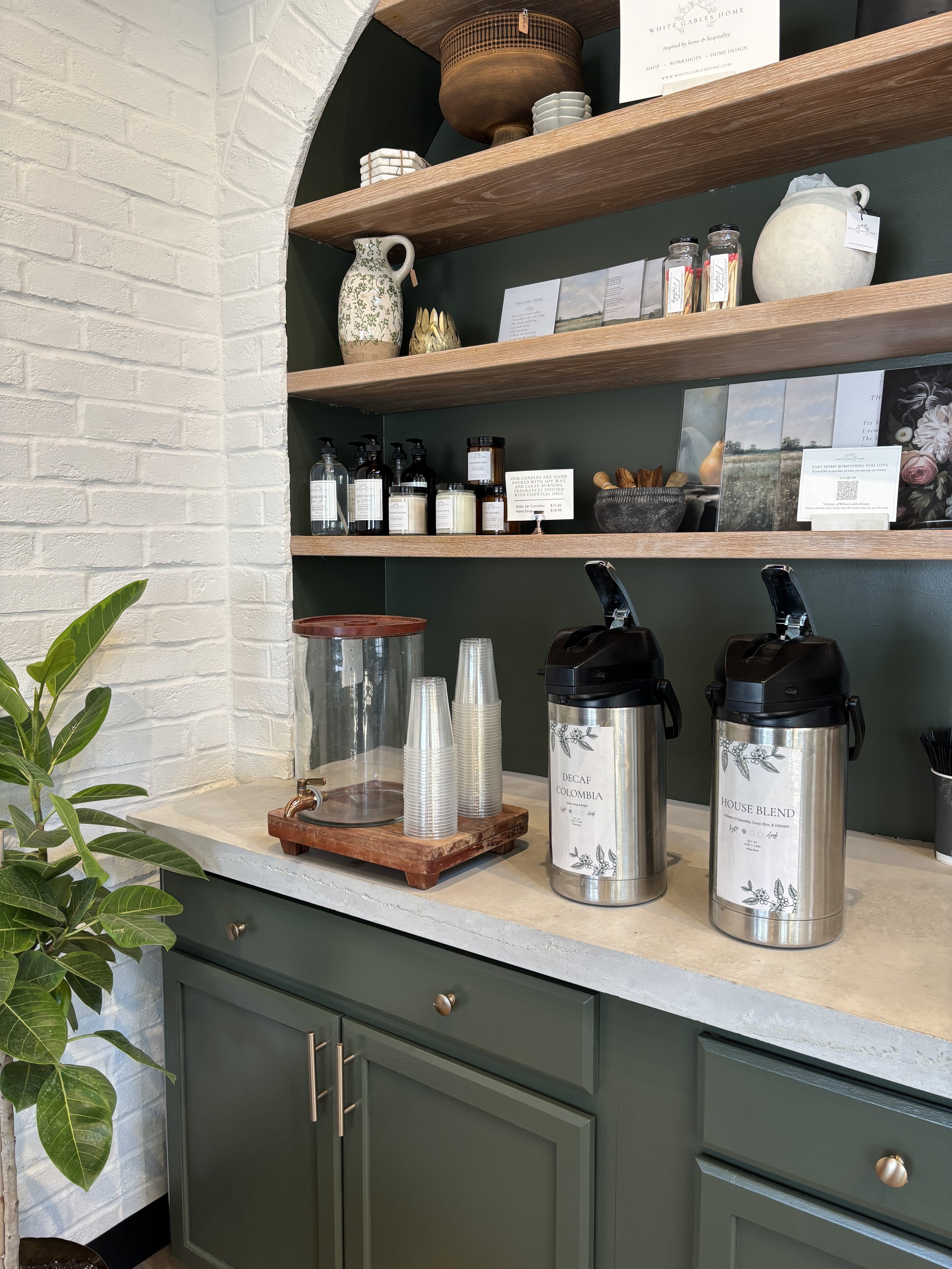 Coffee station with two stainless steel coffee dispensers labeled 'Decaf Colombia' and 'House Blend' on a light-colored counter, with clear cups stacked beside a large glass water dispenser, against a green cabinet and wooden shelves holding ceramic vases, jars, and decor, and a white brick wall on the left side.