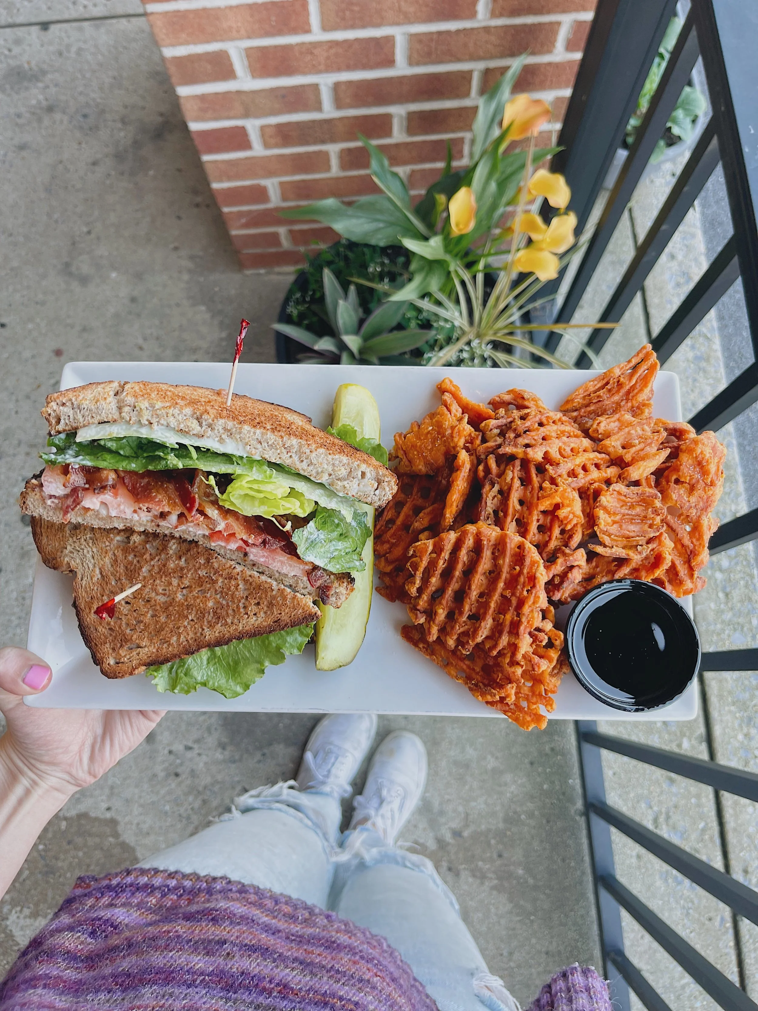 A person holding a white rectangular plate with a sandwich, a serving of pasta, a lime wedge, and a small container of black sauce, standing outdoors near a brick wall with potted plants, wearing a purple sweater and distressed jeans.