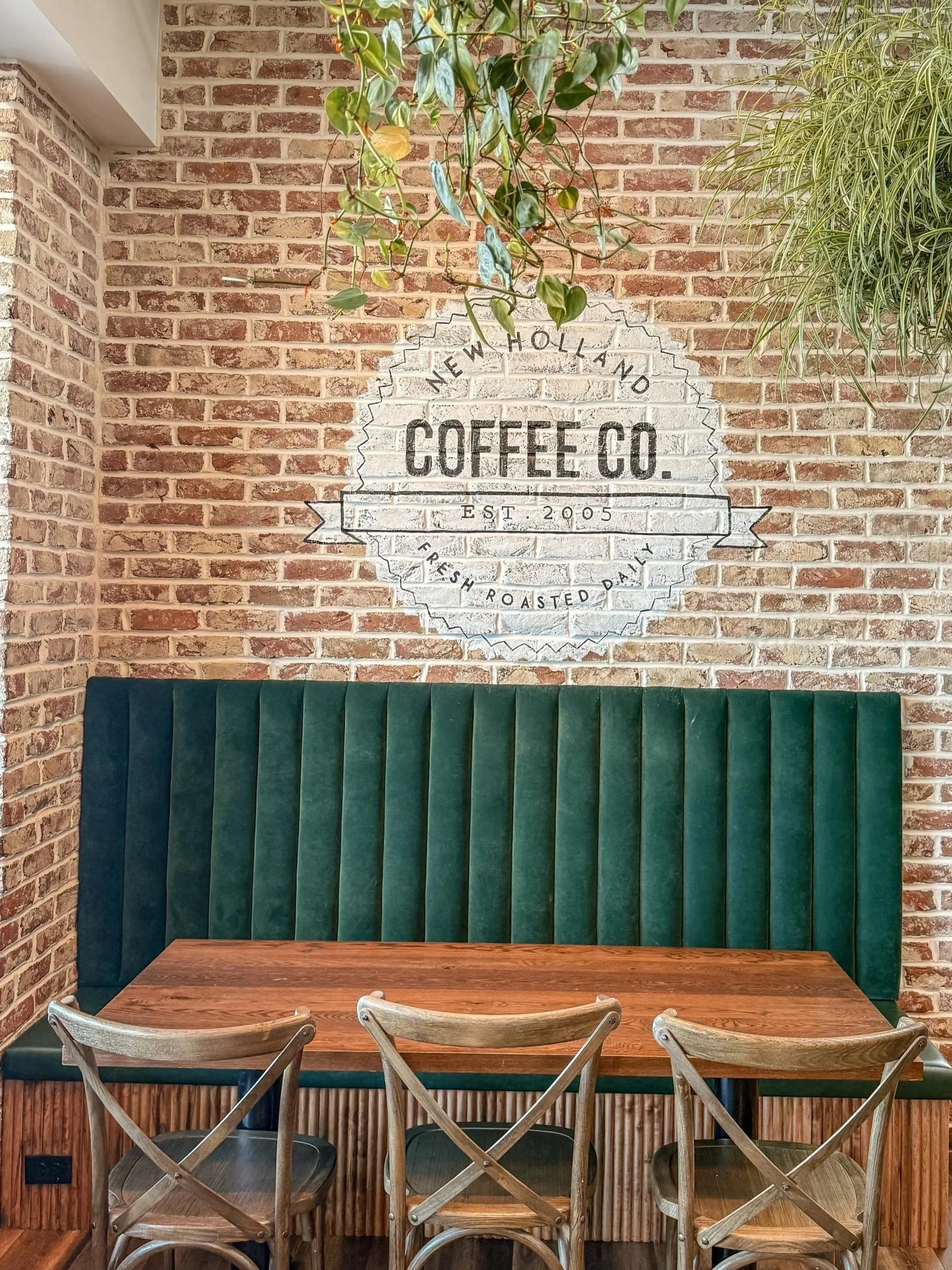 A brick wall with a white painted sign reading "New Holland Coffee Co. Est. 2005" and decorative greenery hanging above the sign. There is a wooden table with four chairs, two wooden and two metal, in a cozy interior setting.