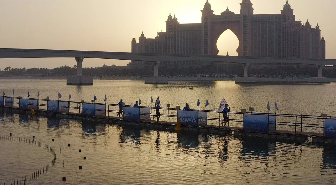 Sunset view of the Atlantis hotel and water park in Dubai with a pedestrian bridge and people walking along a riverside walkway.