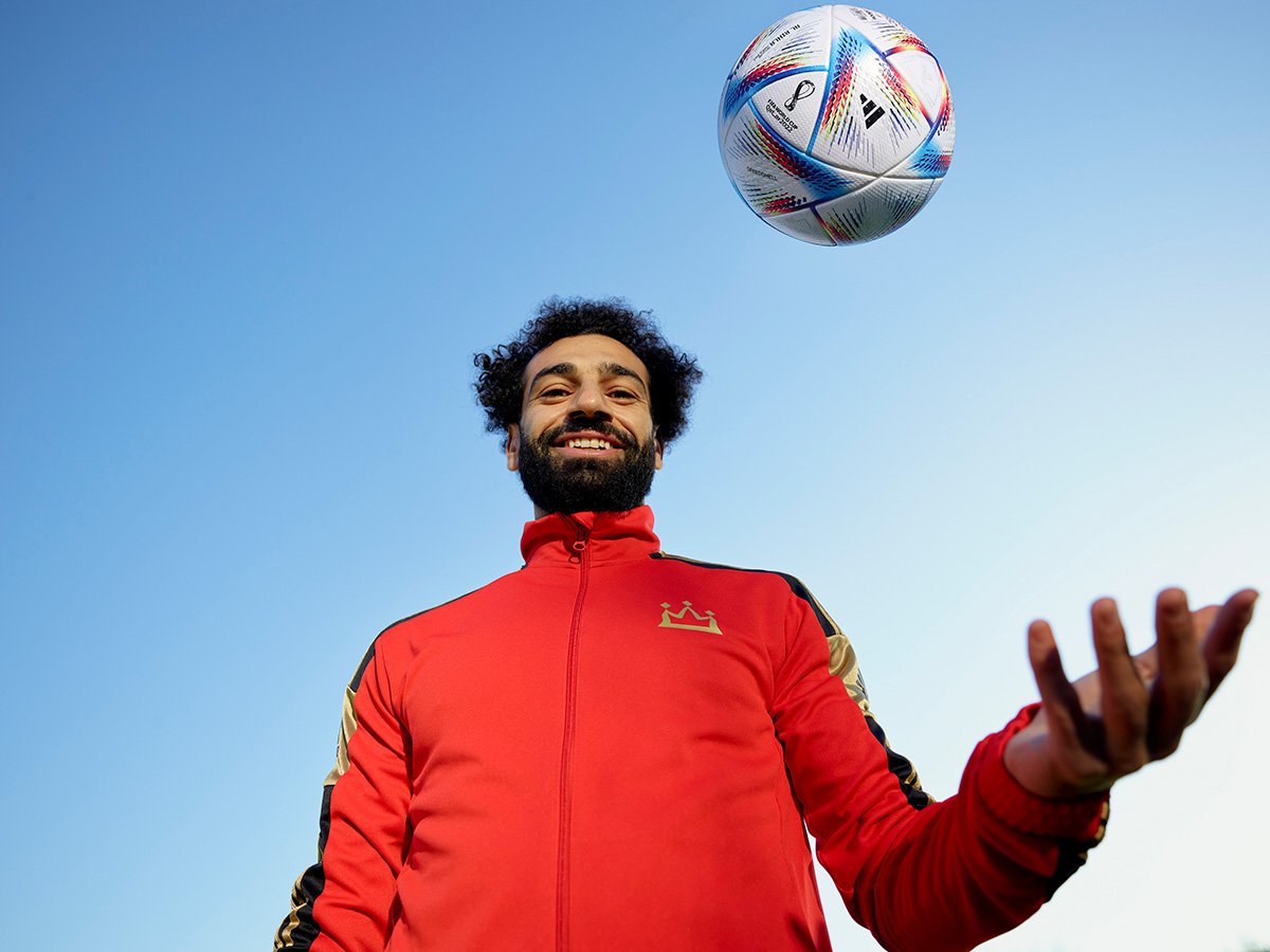 A man with curly hair and a beard, wearing a red sports jacket, smiling while balancing a soccer ball in the air outdoors against a clear blue sky.