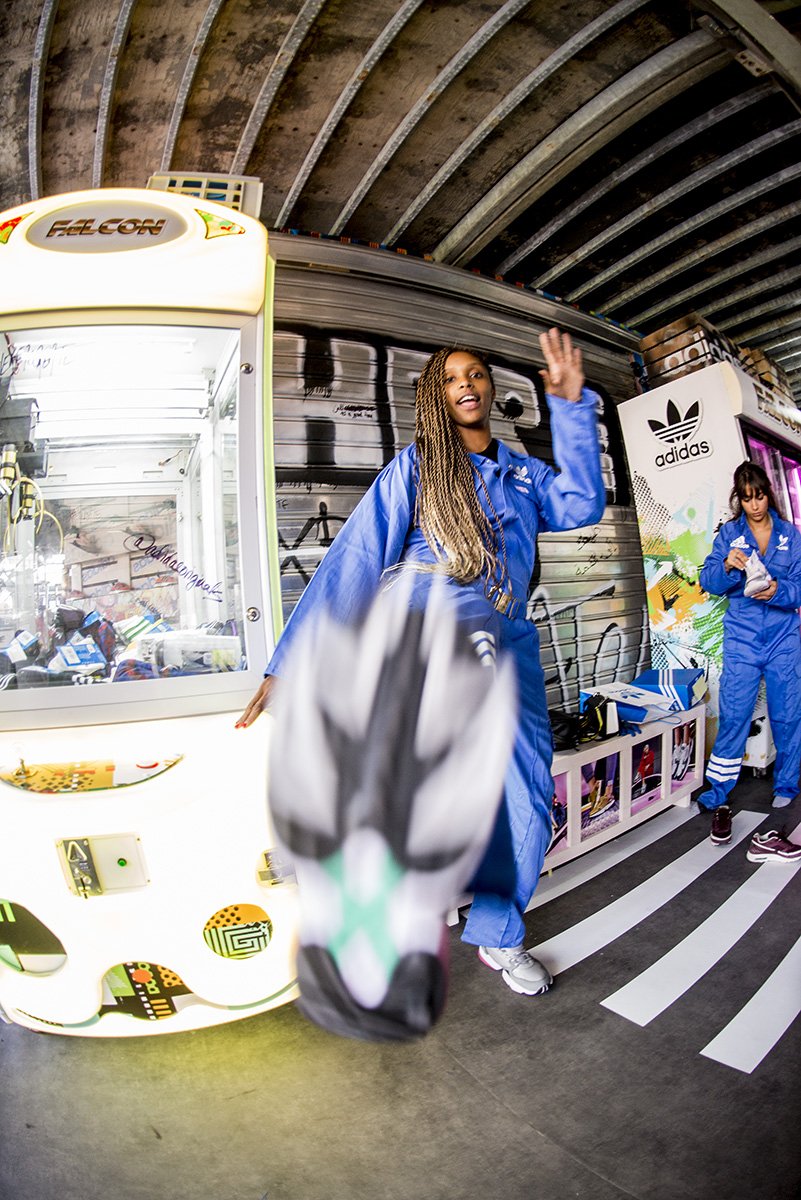 Two women in blue athletic suits near vending machines under a metal arch ceiling. One woman is waving, and a person is holding a sneaker in the foreground.