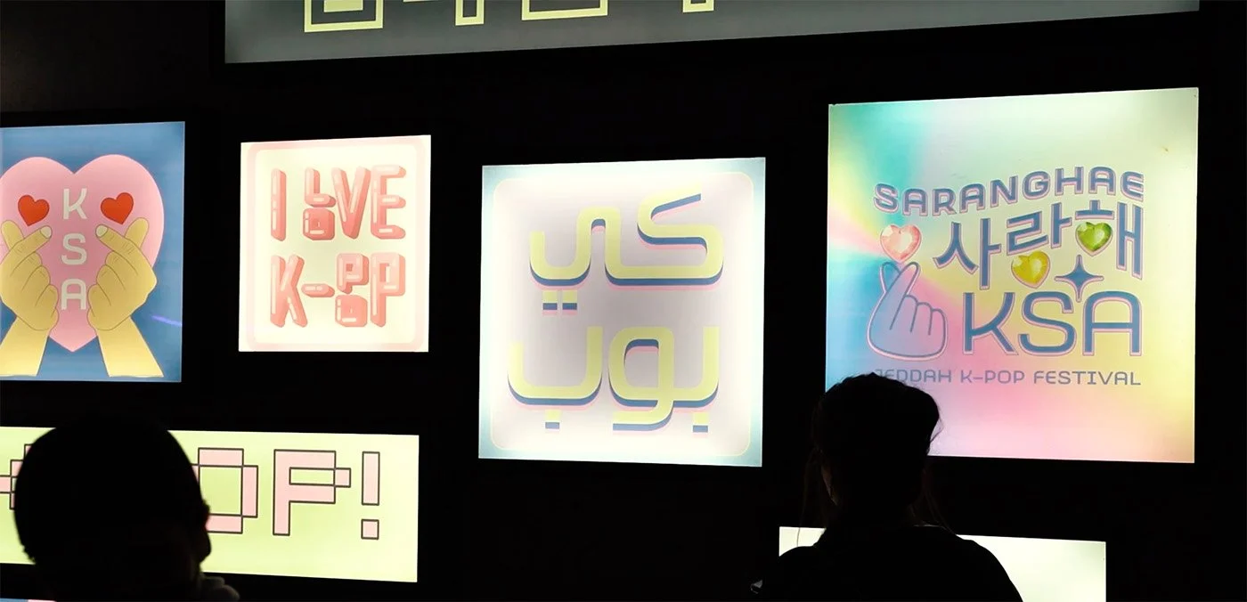 Colorful illuminated posters promoting a K-pop festival, featuring phrases and graphics, with silhouettes of two people in front of the display.