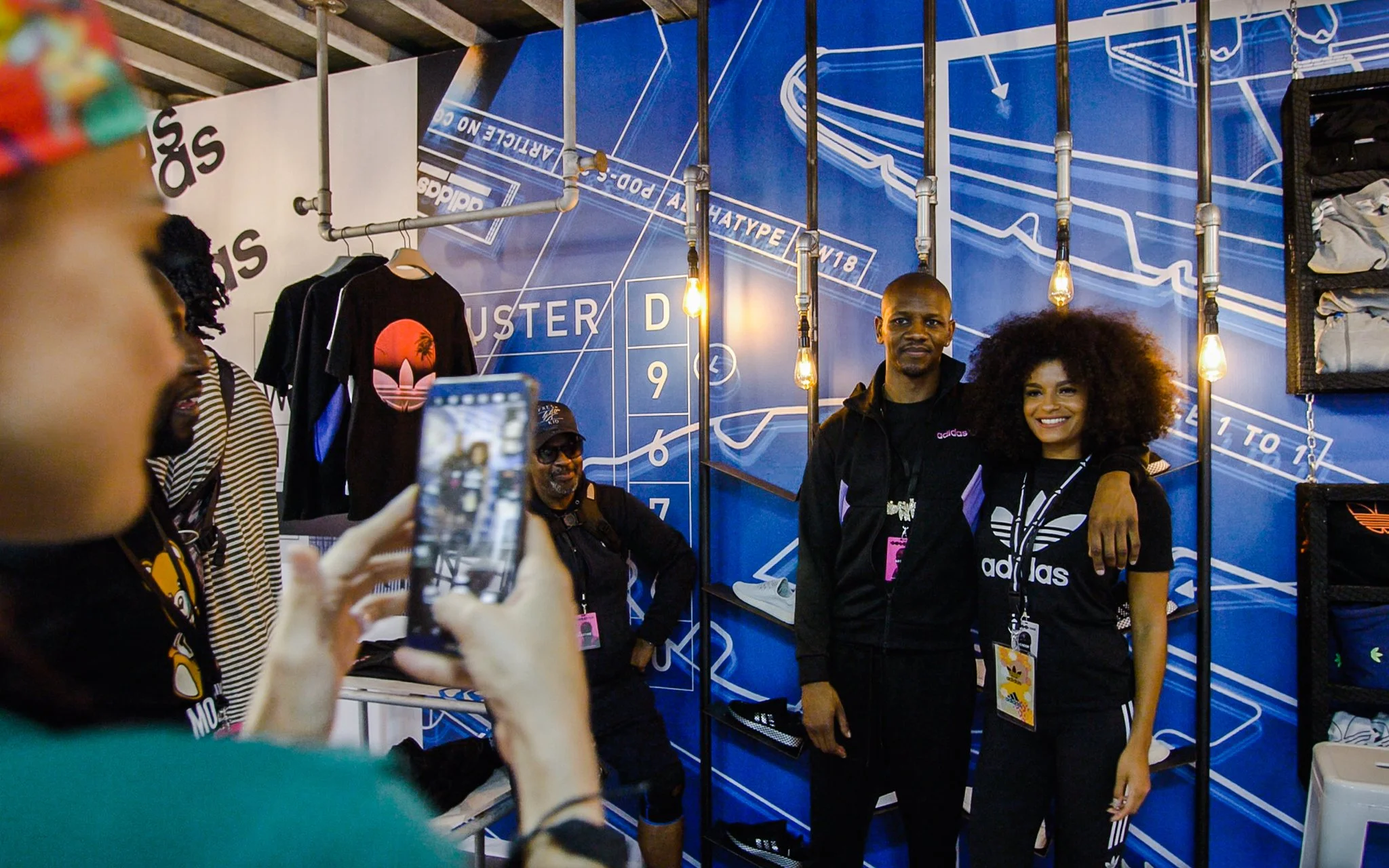 A group of people in black Adidas clothing, with one man and one woman smiling, standing in front of a blue wall with white graphic designs. One person is taking a photo of them with a smartphone. There are hanging light bulbs and clothing items on display.