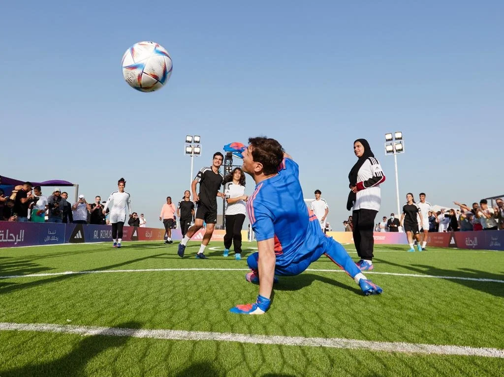 A soccer match showing a goalkeeper in blue uniform performing a bicycle kick during the game on a green field with a crowd watching on, under a clear blue sky.