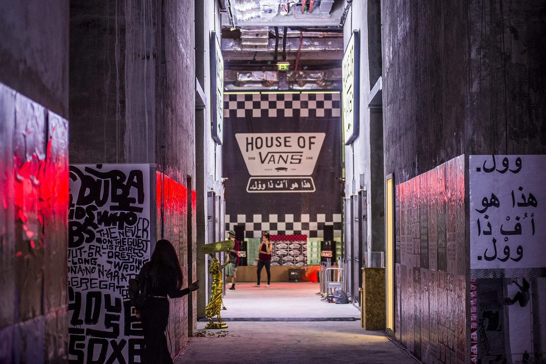 Indoor urban space with a large sign reading 'House of Vans' at the end of a corridor surrounded by posters and artwork, with a few people walking and talking.