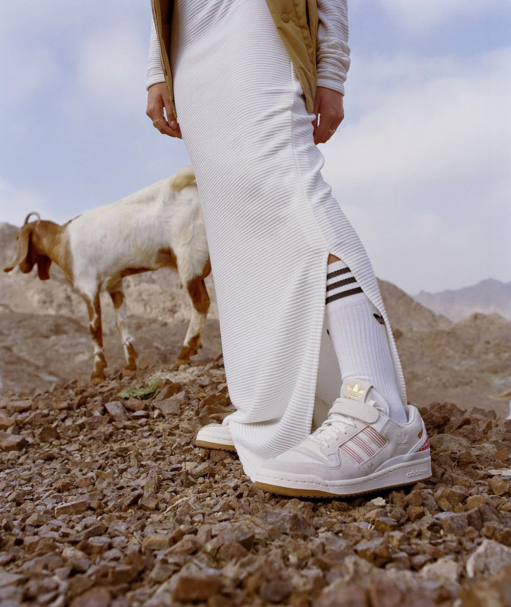 Person wearing white Adidas sneakers, white ribbed skirt, and striped socks standing on rocky terrain in a desert landscape with a goat nearby and mountains in the background.