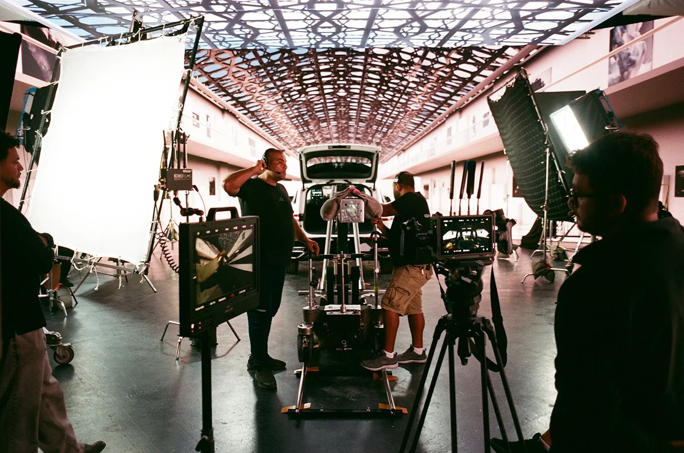 Film crew setting up equipment in a spacious indoor studio with large lights and a camera on a dolly. Crew members are adjusting equipment, preparing for a shoot, with a vehicle in the background and decorative ceiling panels.
