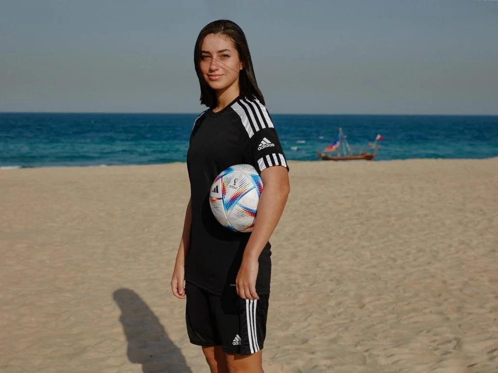 A young woman in a black Adidas sports outfit stands on a sandy beach, holding a colorful volleyball under her arm, with the ocean and a small boat in the background.