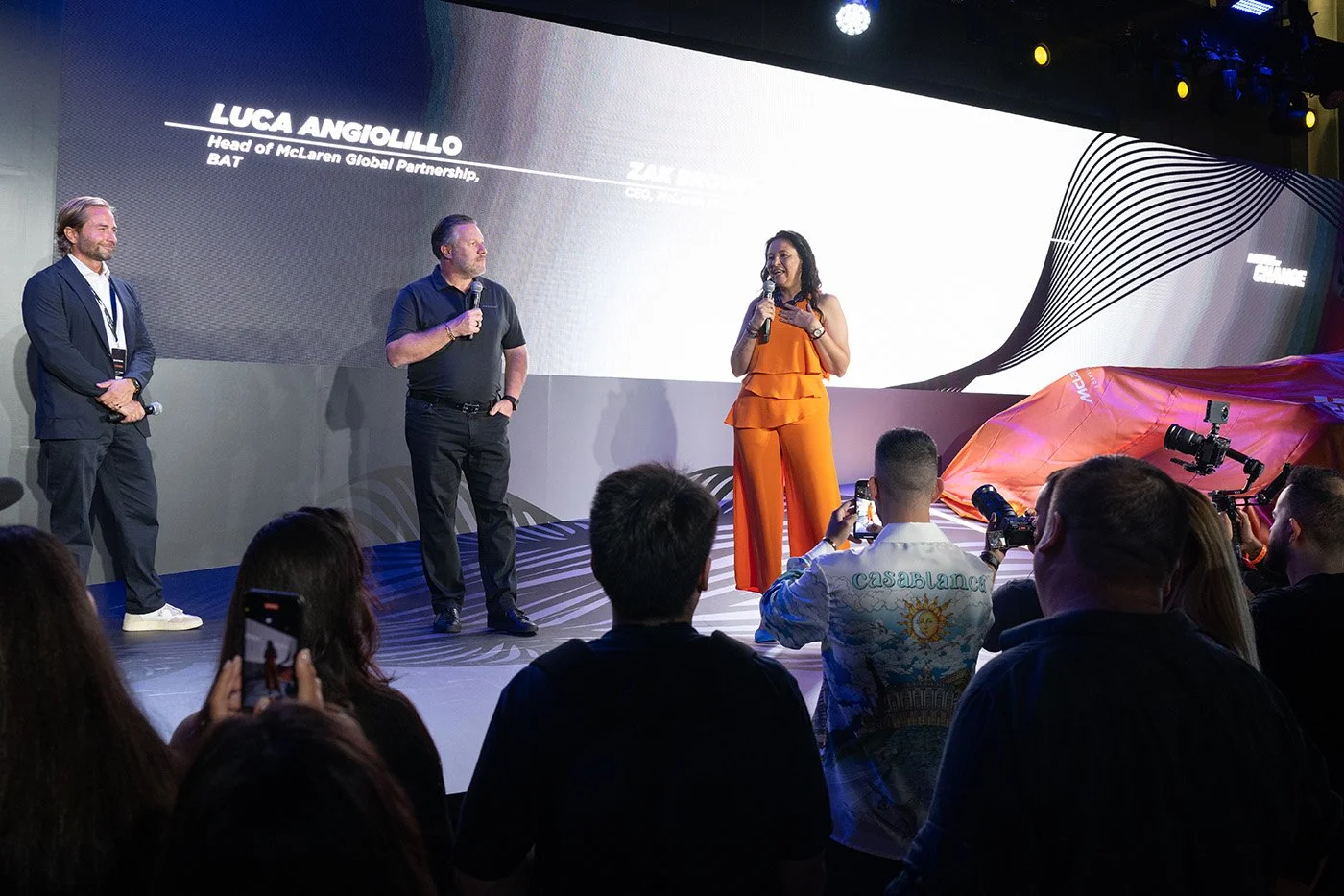 Three people on stage during a presentation or event with a large screen behind them. The woman in orange is speaking into a microphone. The man in the middle is also holding a microphone, and the man on the left is standing with his hands clasped. Audience members are visible, some taking photos.