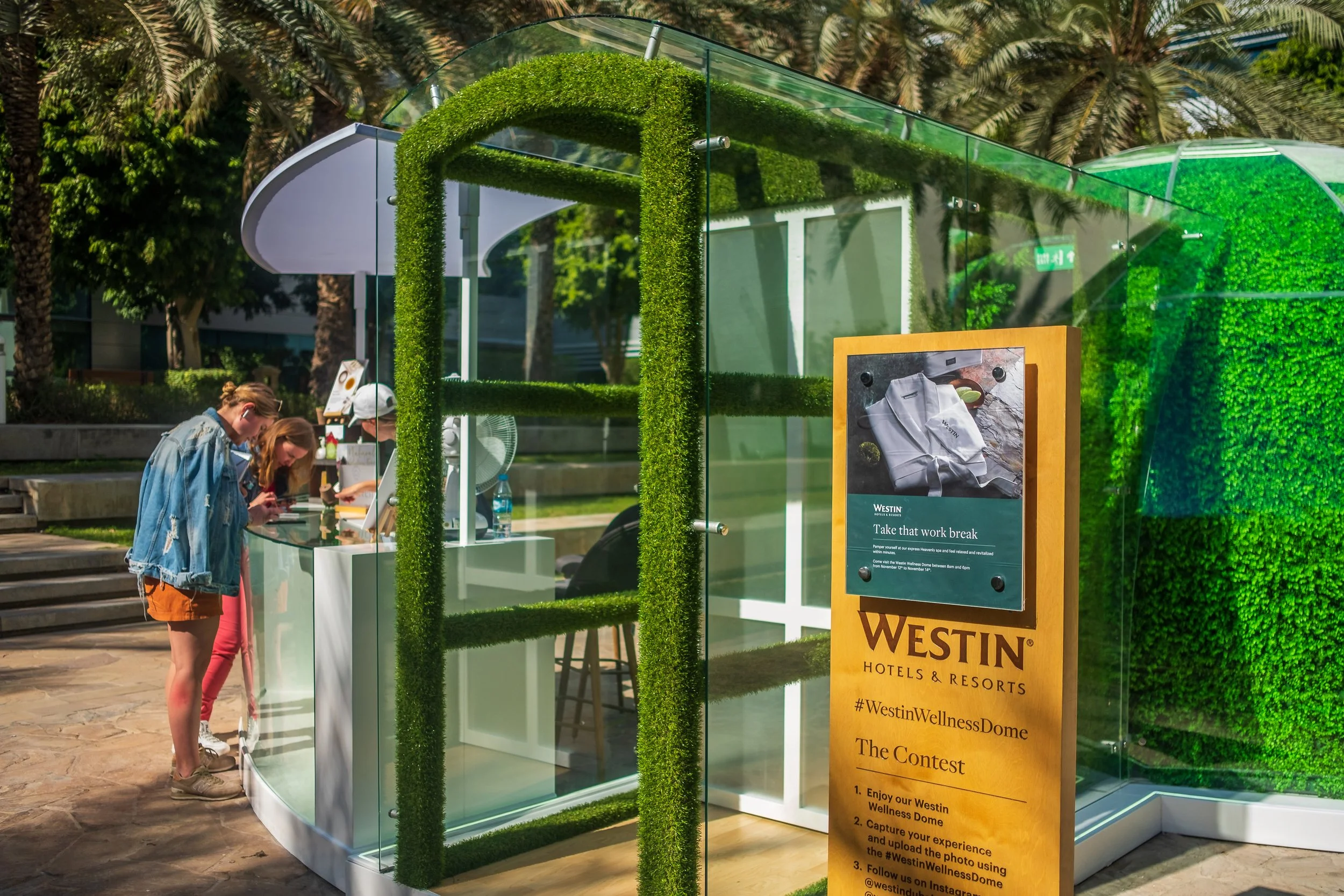 Two girls looking at a glass display booth with greenery accents in an outdoor setting. A sign for Westin Hotels & Resorts is visible, promoting a photo contest at the wellness dome.