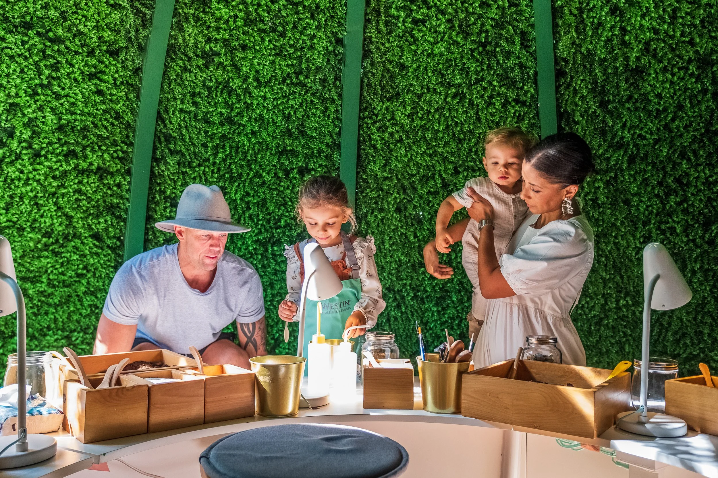 Family gathered at a table in front of a green leafy wall, with night light illuminating the scene, engaged in an activity, possibly crafting or baking.