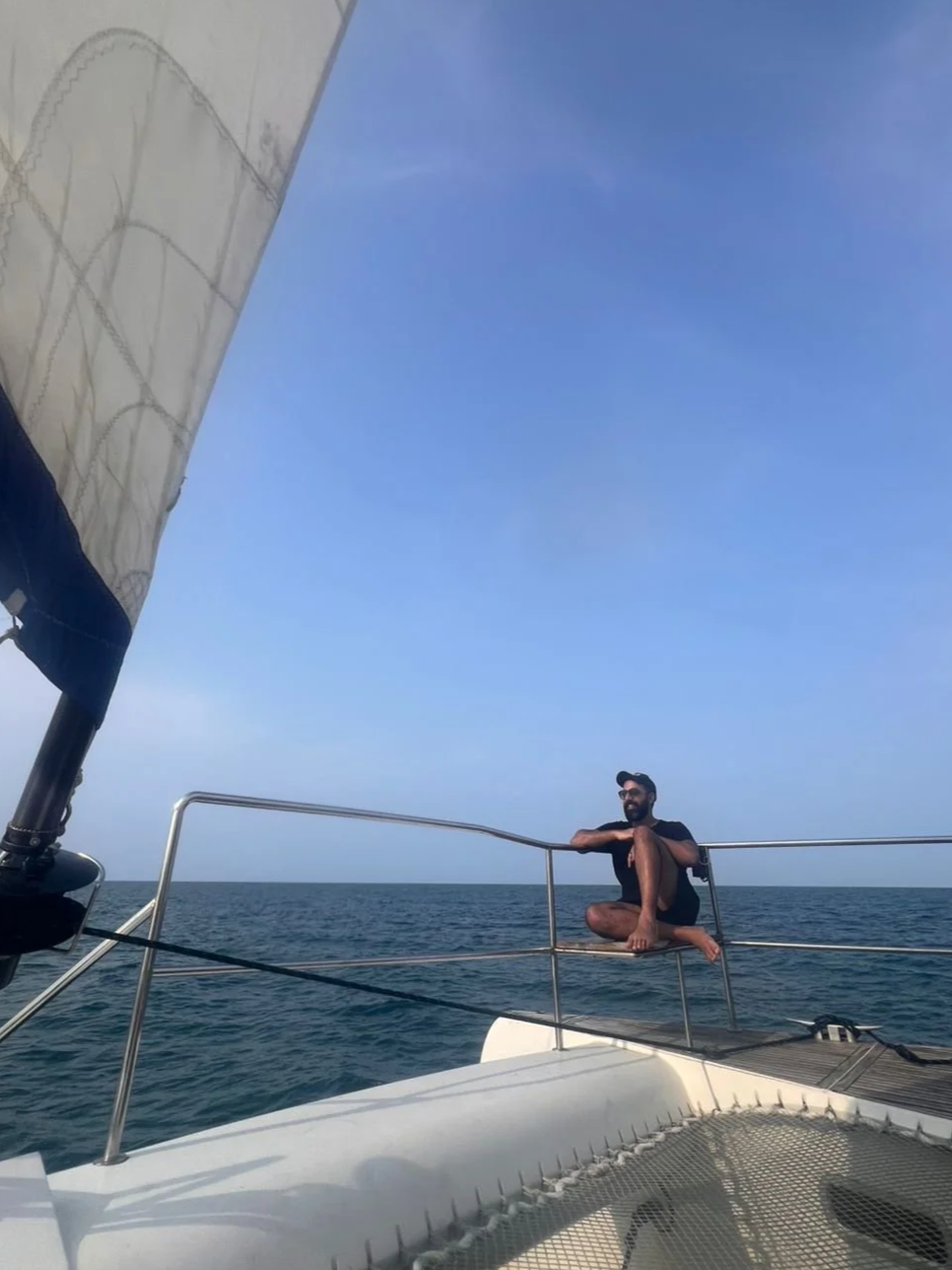 A man sitting on the edge of a sailboat's bow, smiling and relaxing while surrounded by open water and clear blue sky.