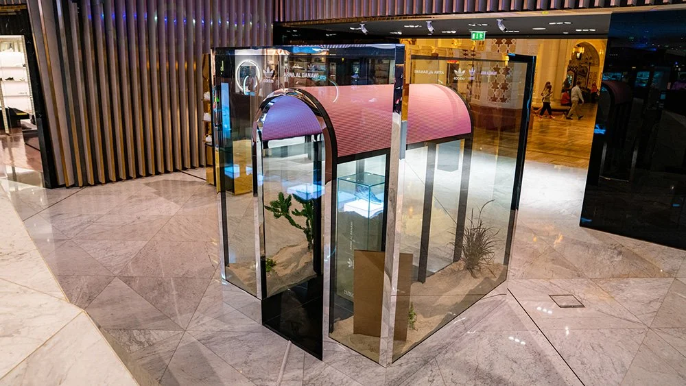 Decorative glass enclosure with desert plants and rocks, featuring a pink curved canopy, situated inside a shopping mall.